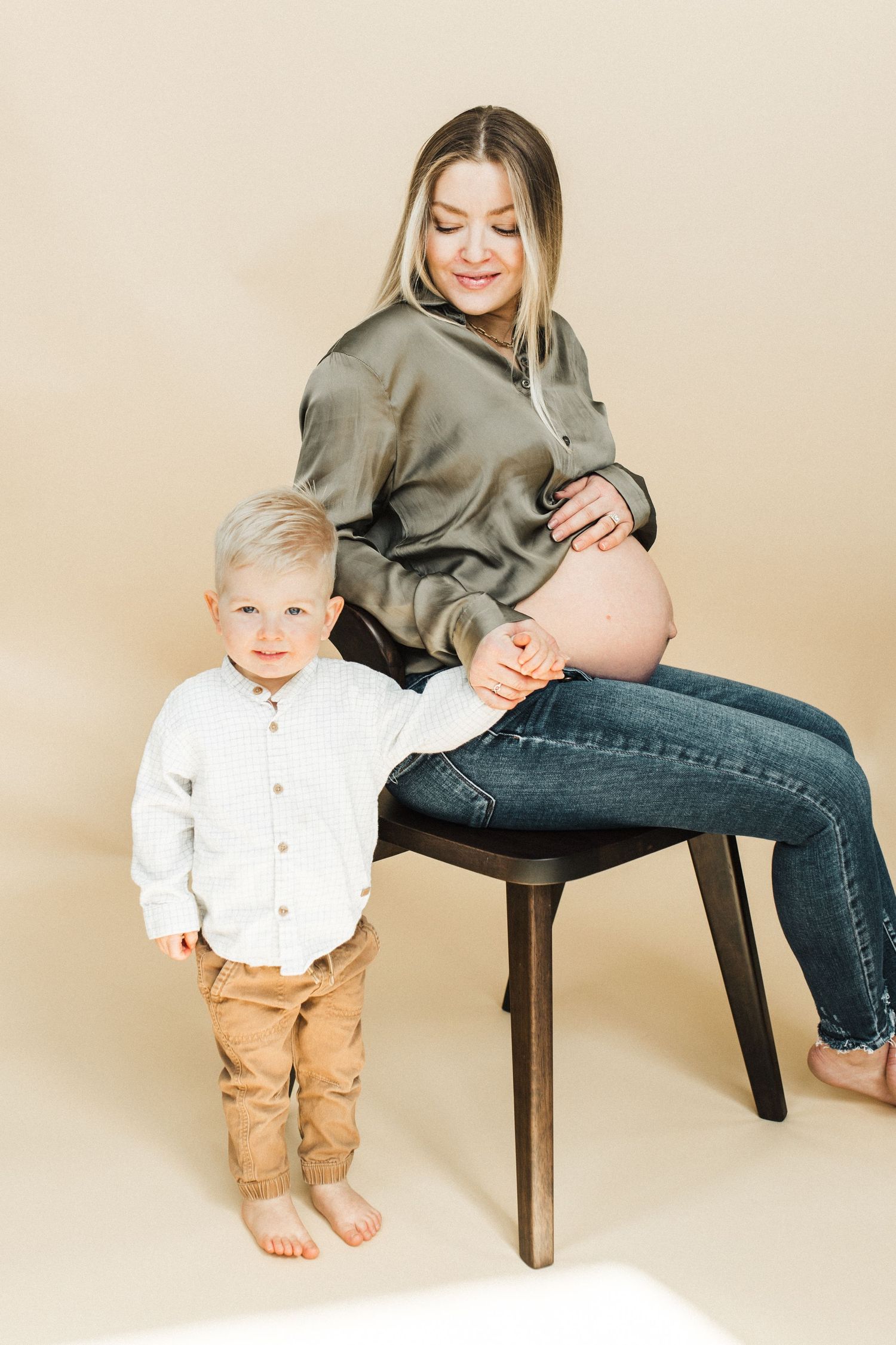 Maternity photo featuring toddler standing beside pregnant mother on chair.