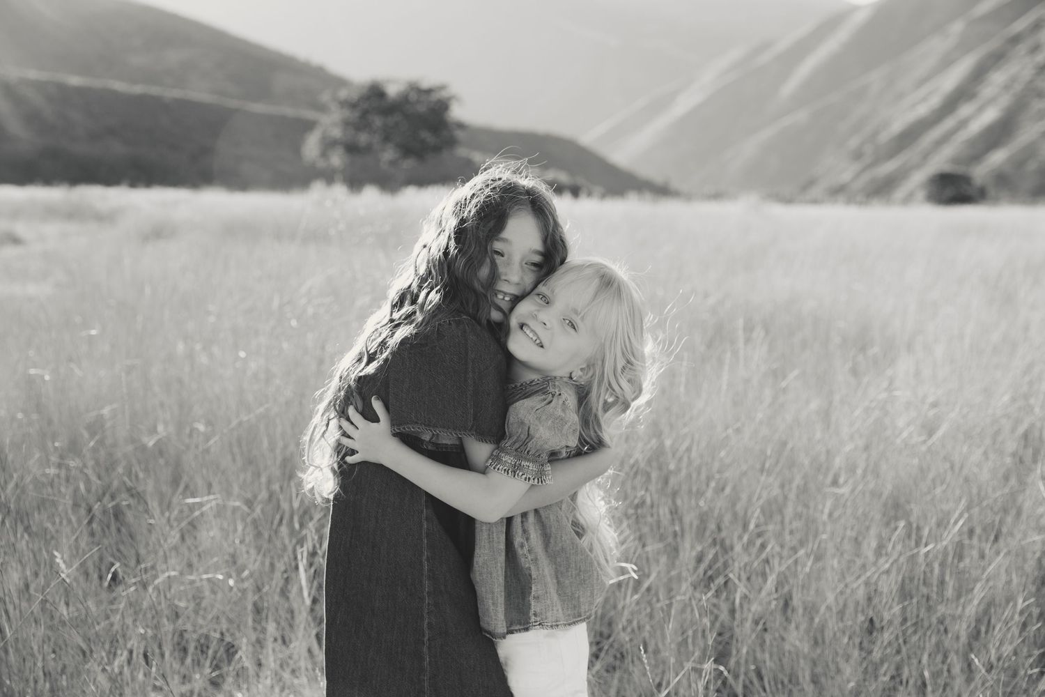 A couple embraces in a sunlit meadow with mountains in the background, captured in black and white.