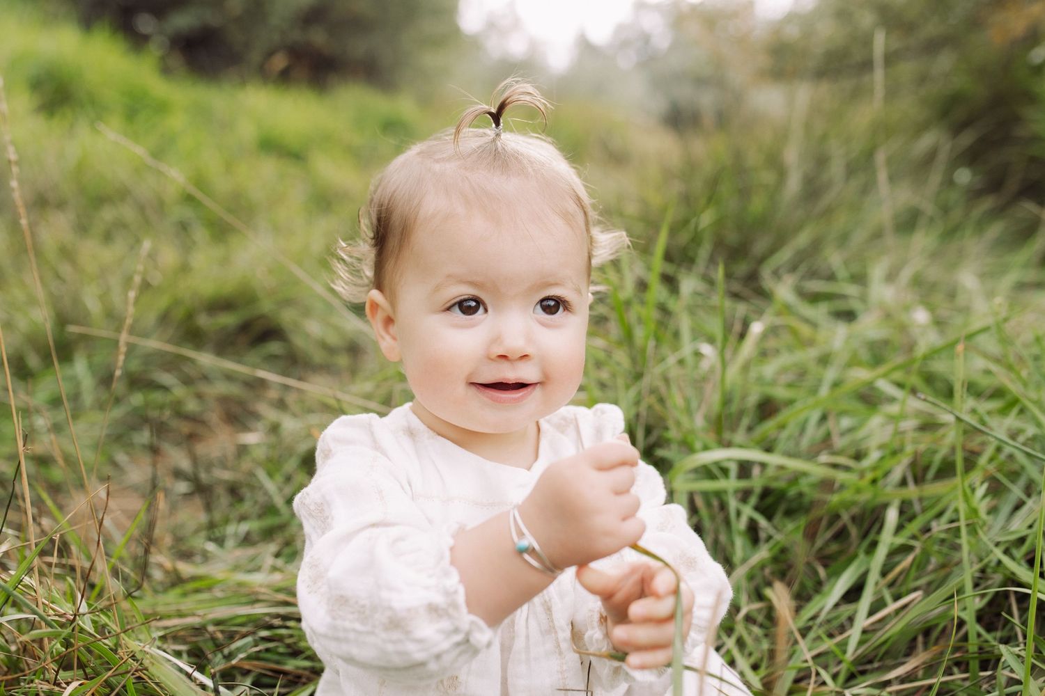 A toddler in a white dress plays in tall grass on a sunny day.