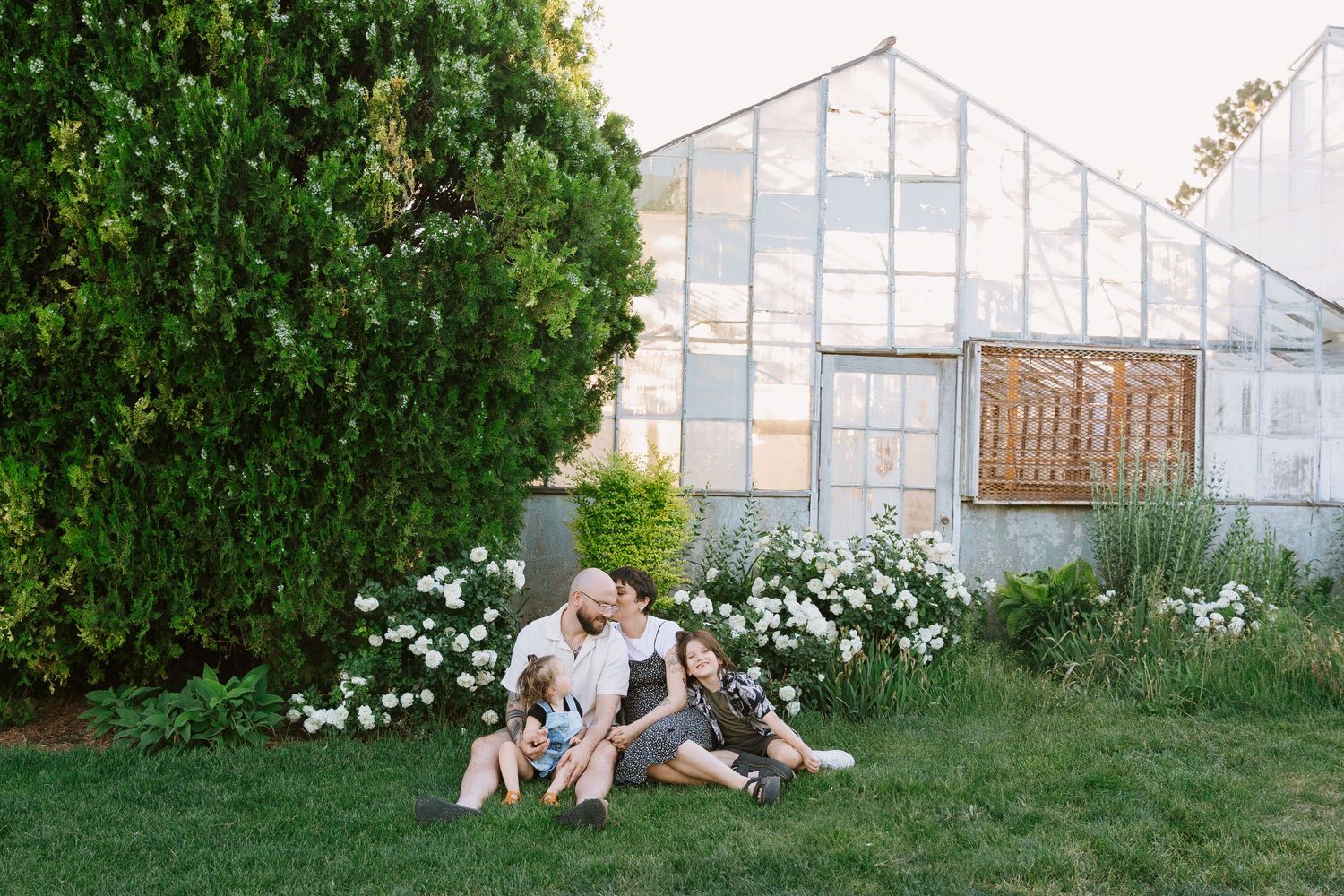 A couple sits together on a grassy lawn beside a greenhouse in a romantic outdoor setting.