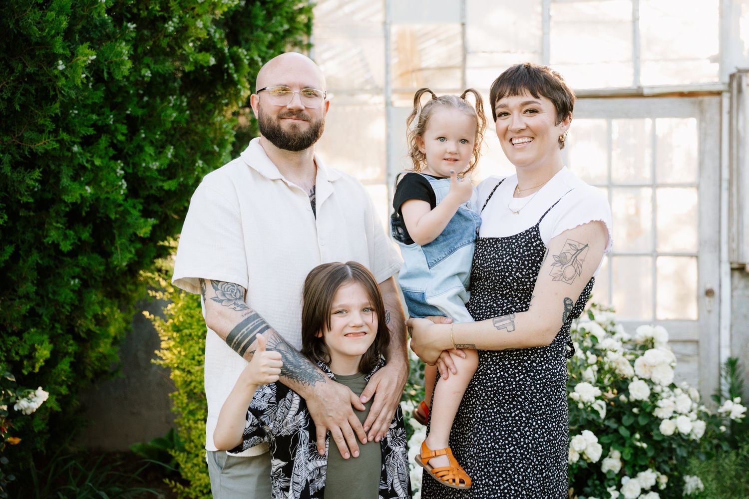 A family poses together outdoors against a wall with greenery and white flowers in the background.