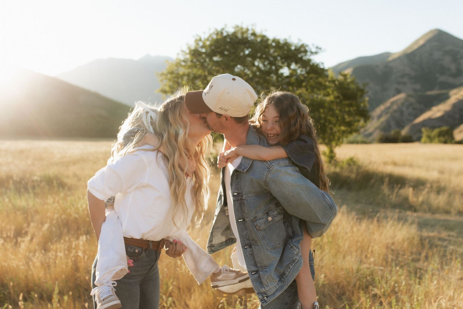 A group embraces while standing in a sunlit meadow with mountains visible in the distance.