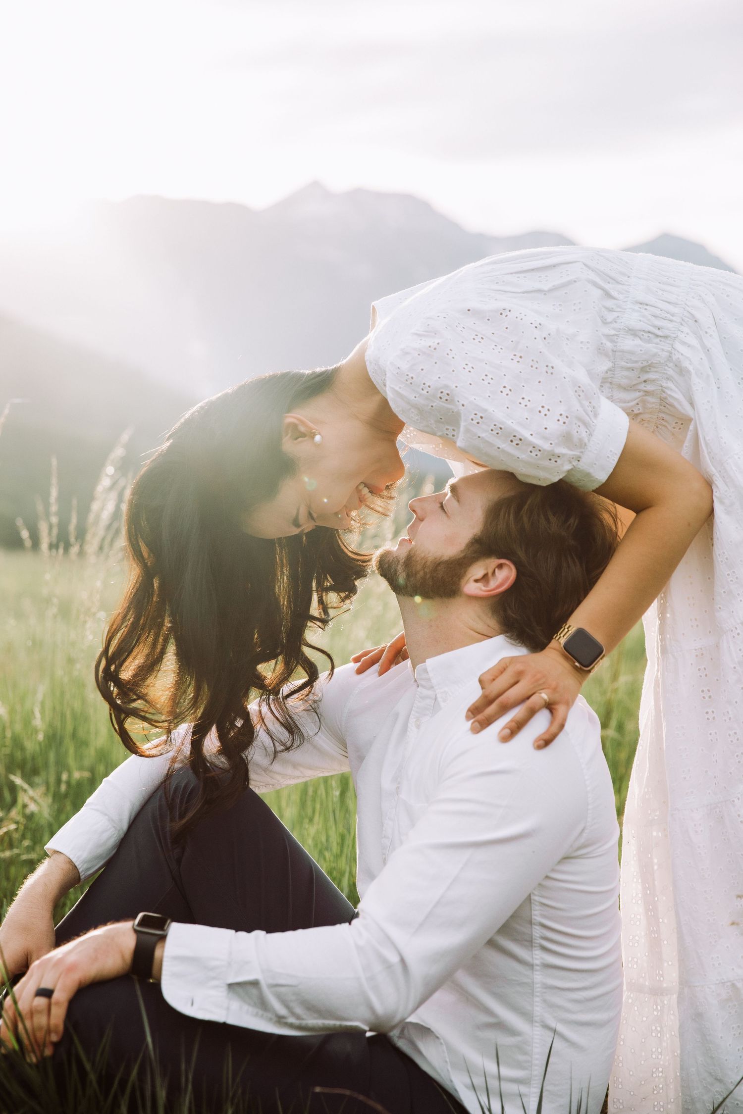Playful couple in white clothing sharing intimate moment in mountain field.