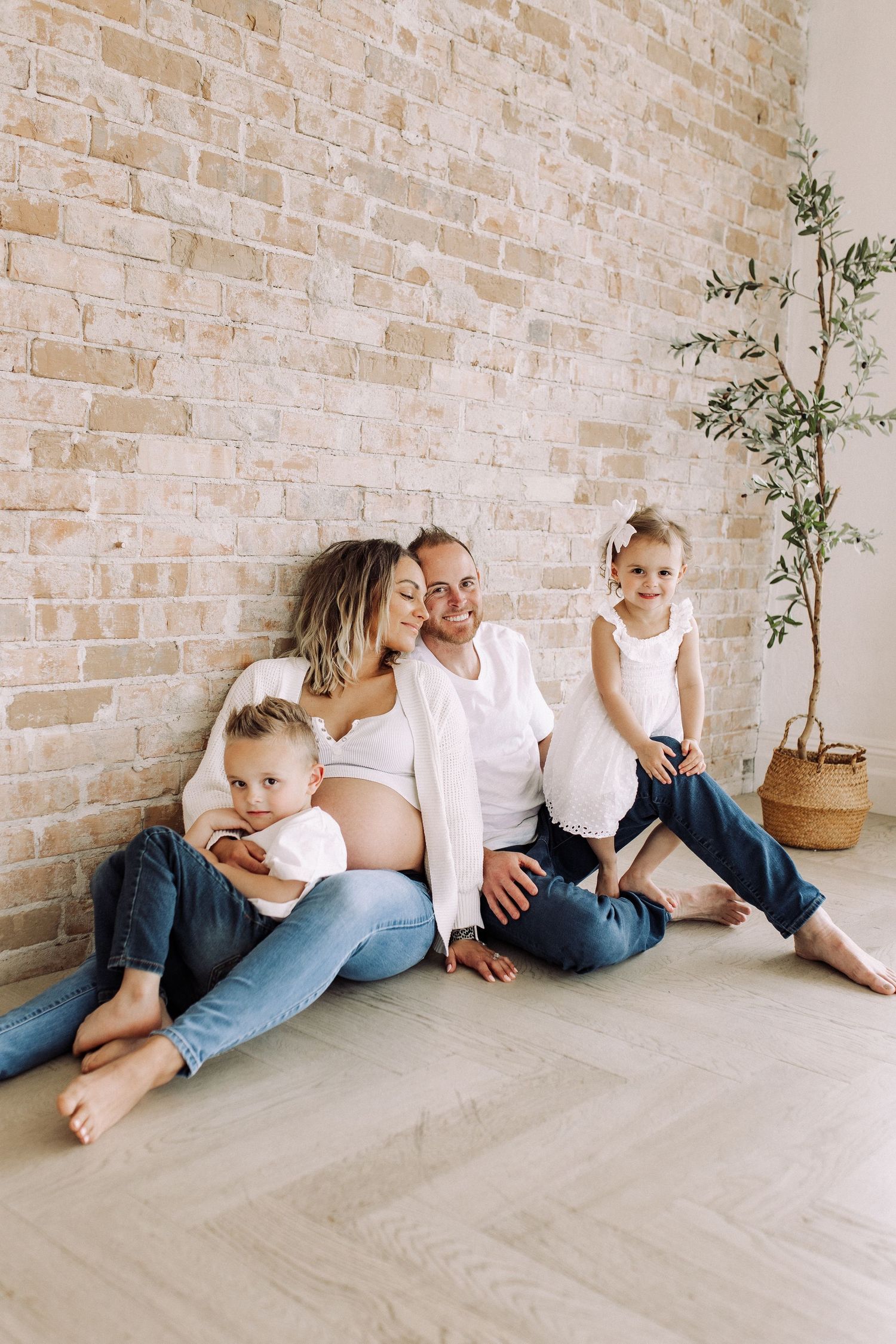 Family sitting together on floor against brick wall during maternity session.