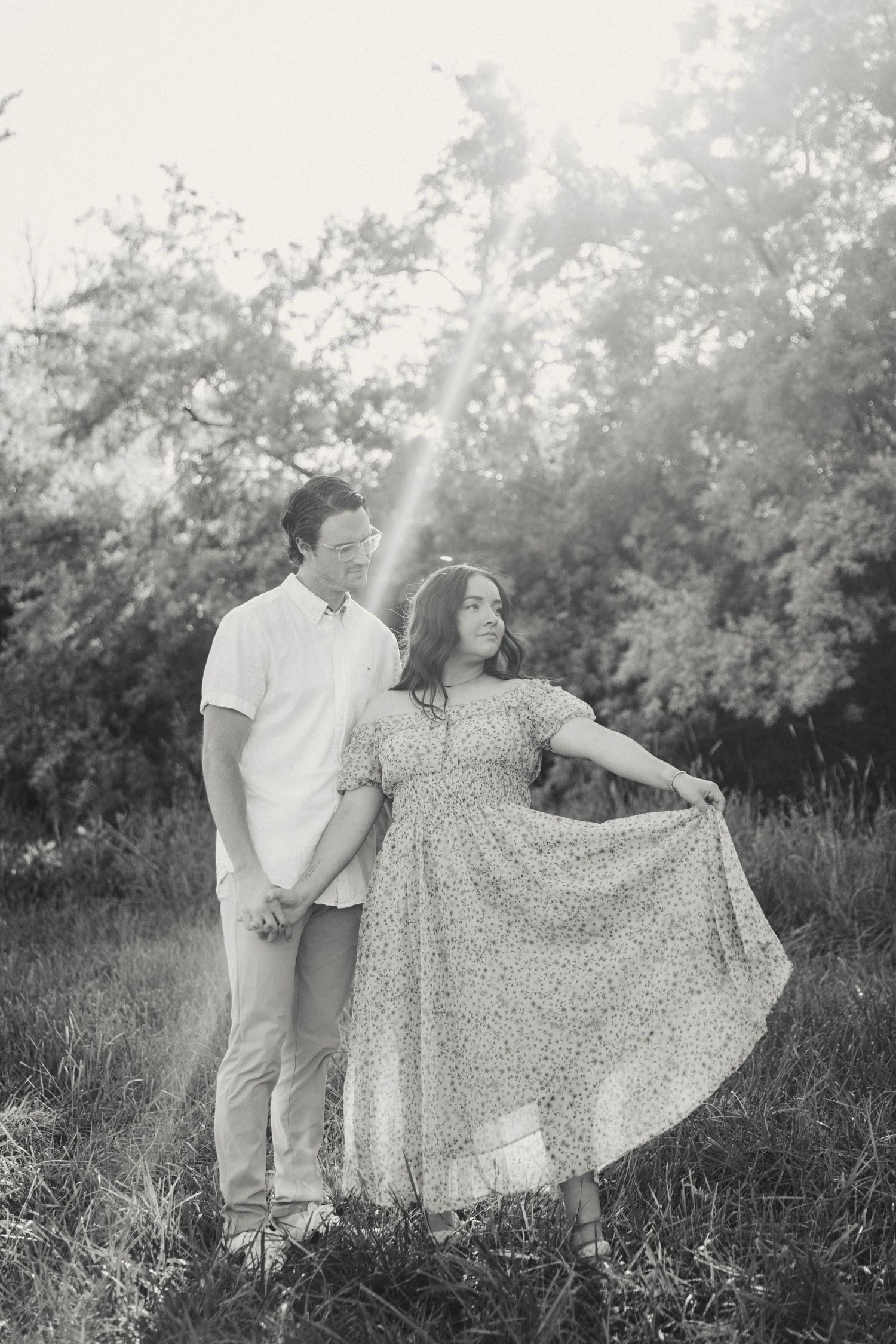 A couple poses in a meadow as sunlight filters through trees, with her floral dress flowing in the breeze.