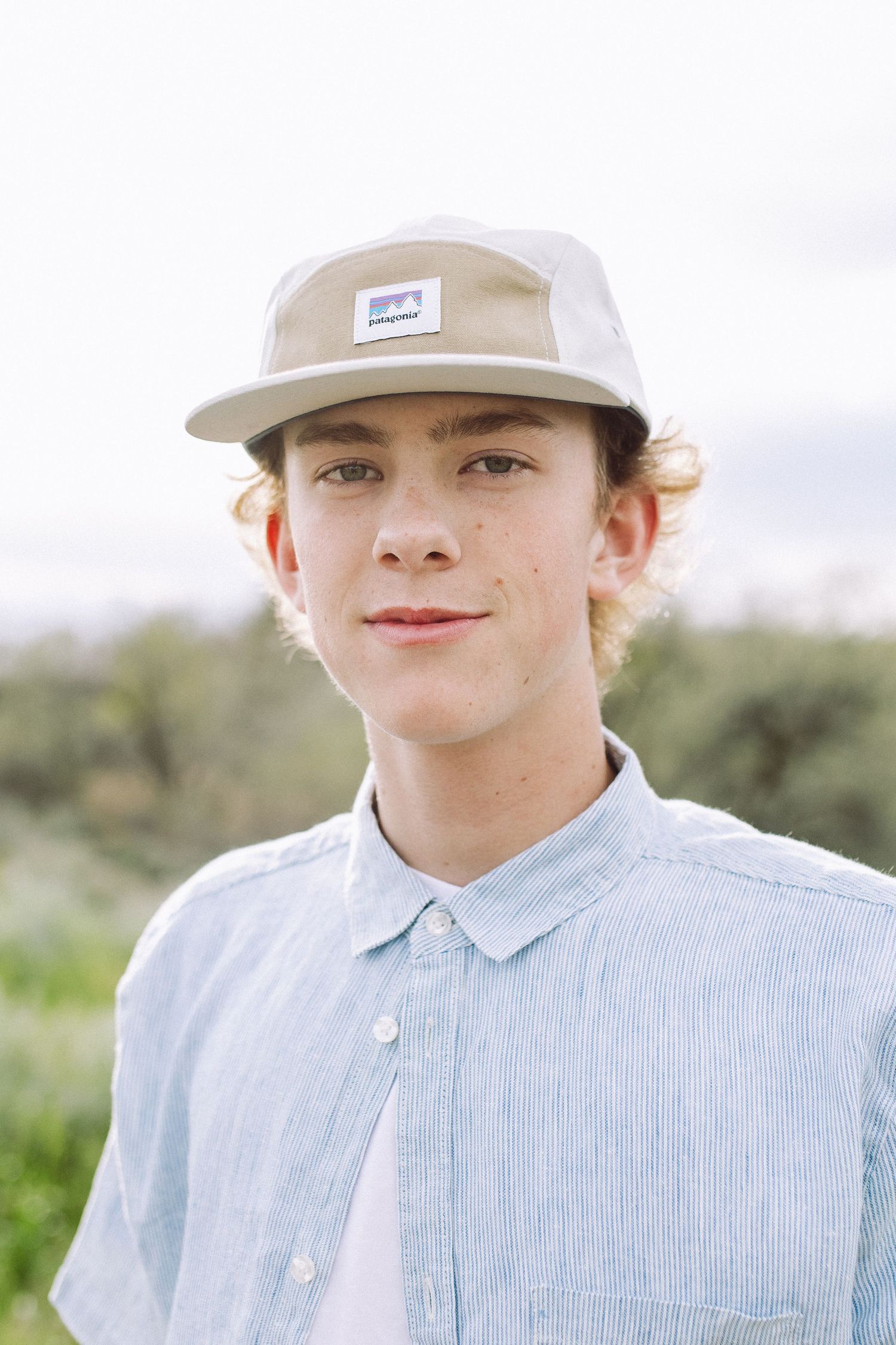 Portrait in light blue shirt and beige cap against natural background.
