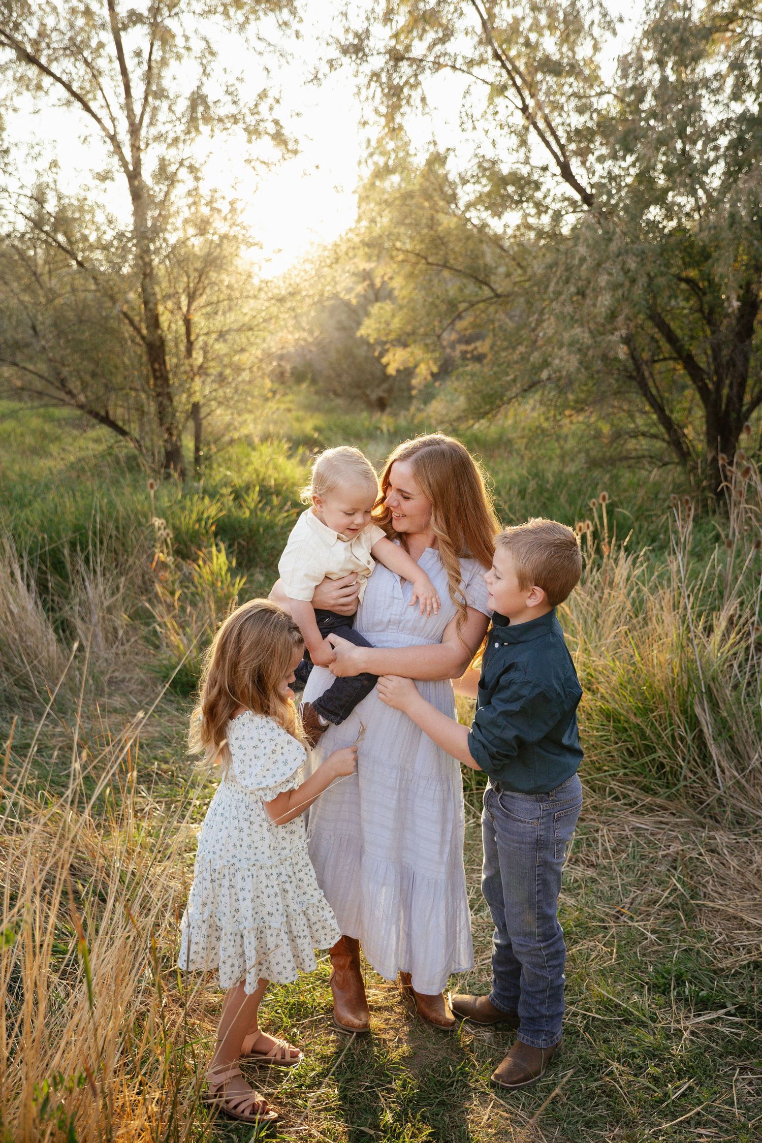 A family shares a tender moment together in a sun-drenched field with tall grass and trees during golden hour.