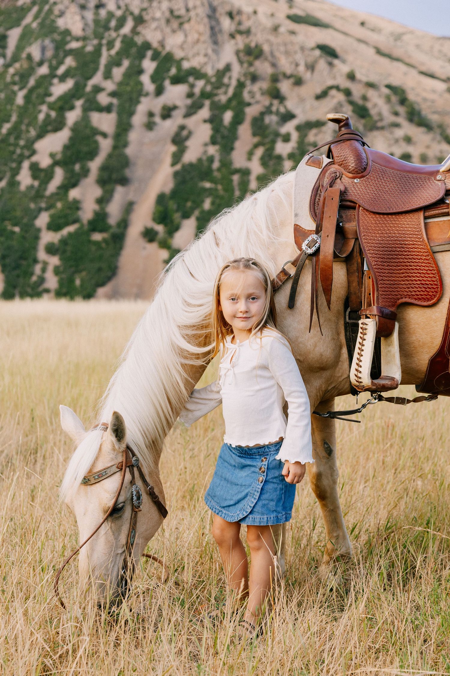 Children play and interact with a white horse in a grassy field with mountains in the background.