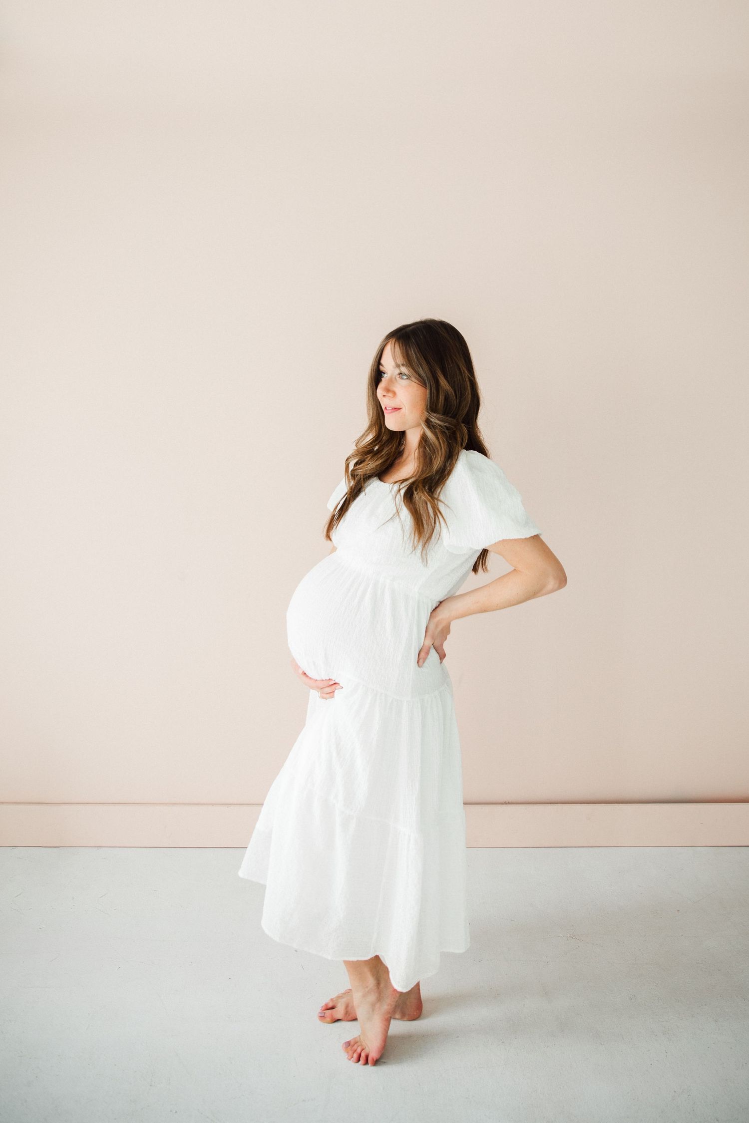 Pregnant woman in flowing white dress stands barefoot against beige wall.