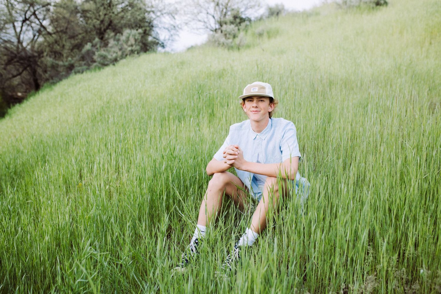 Figure seated in lush green hillside wearing casual summer clothing.