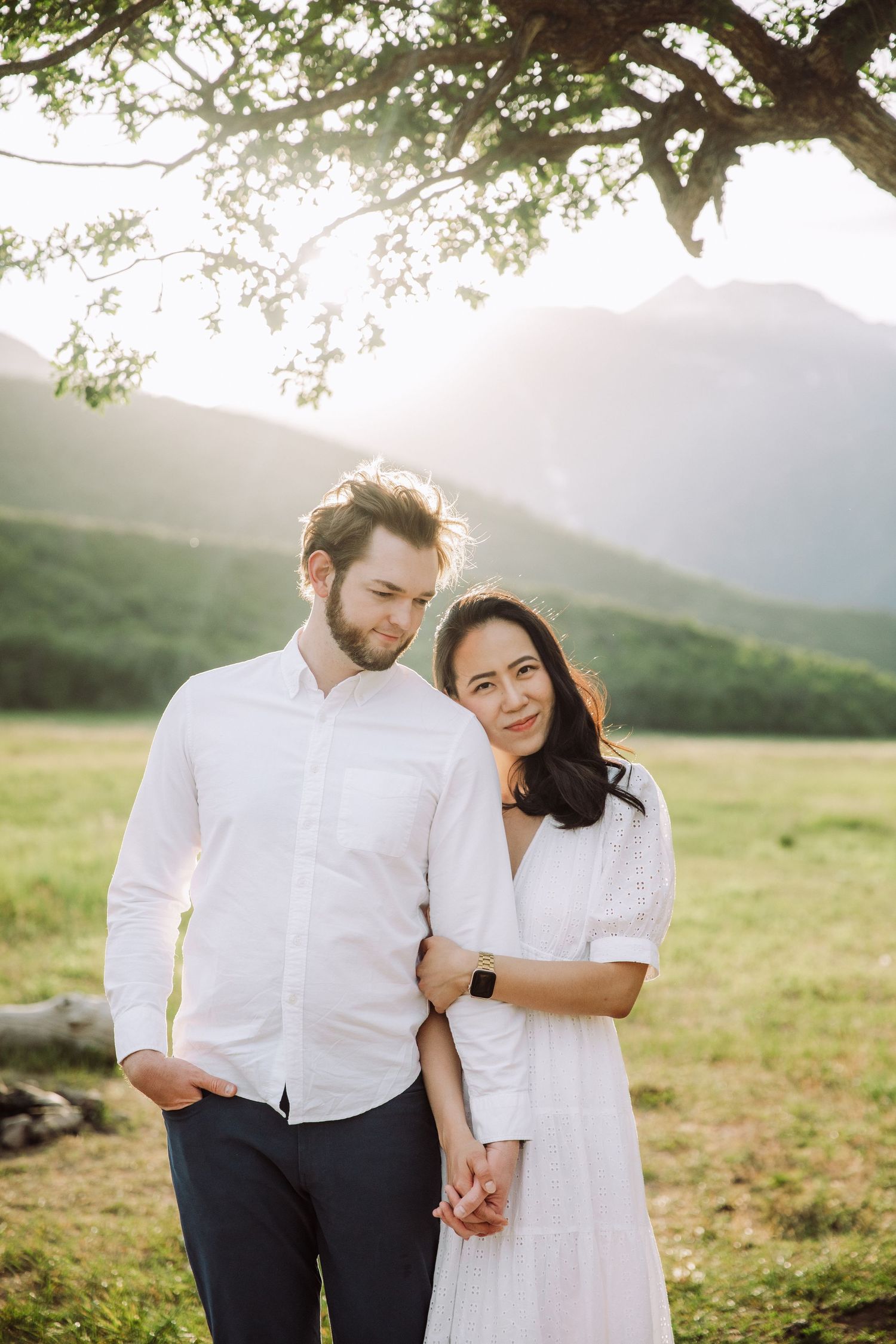 Couple in white clothing pose together under golden sunlight with mountains in background.