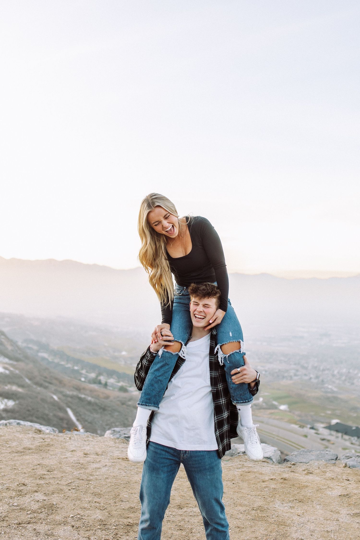 Two people share a playful moment on a mountain overlook wearing casual attire.