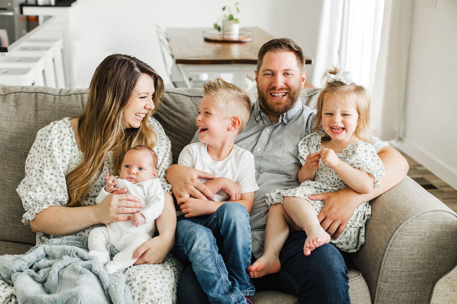 A family laughing together while relaxing on a grey couch in a bright living room.