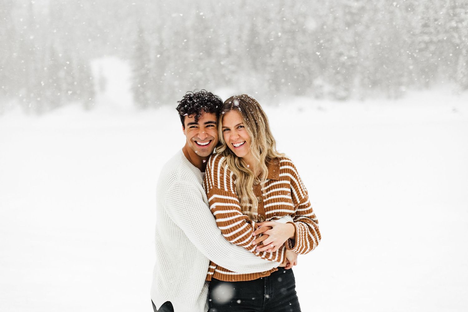 A couple in matching striped sweaters smiling together in a snowy winter landscape.
