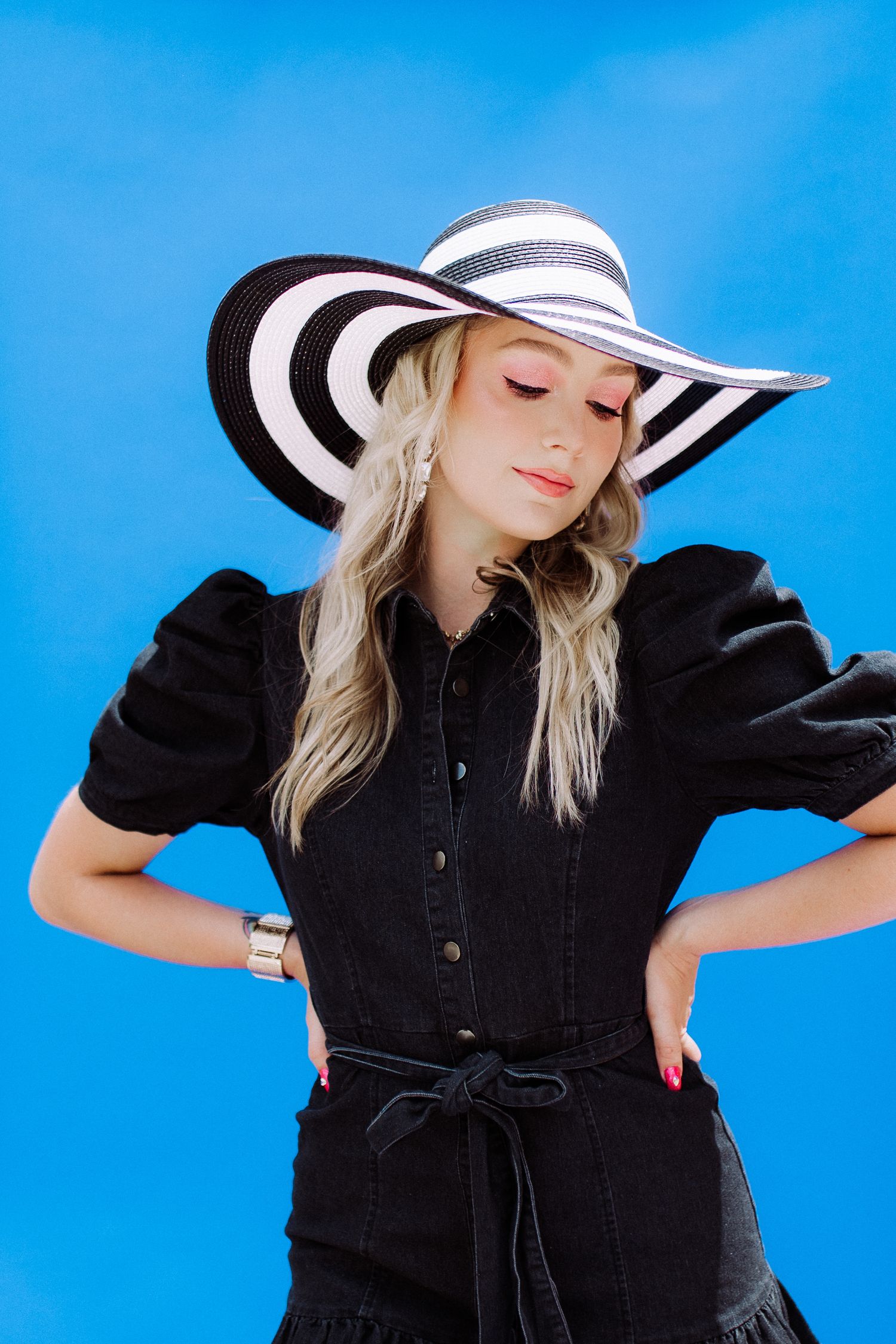 Fashion model wearing black denim dress and striped sunhat poses dramatically against blue backdrop.