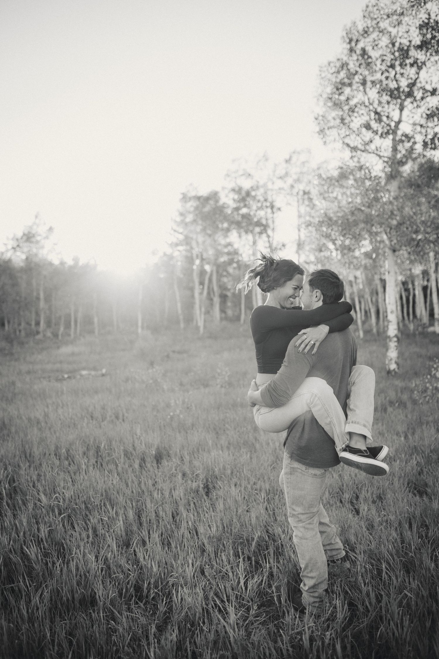 A black and white photo of a couple embracing in a sunlit meadow surrounded by trees.