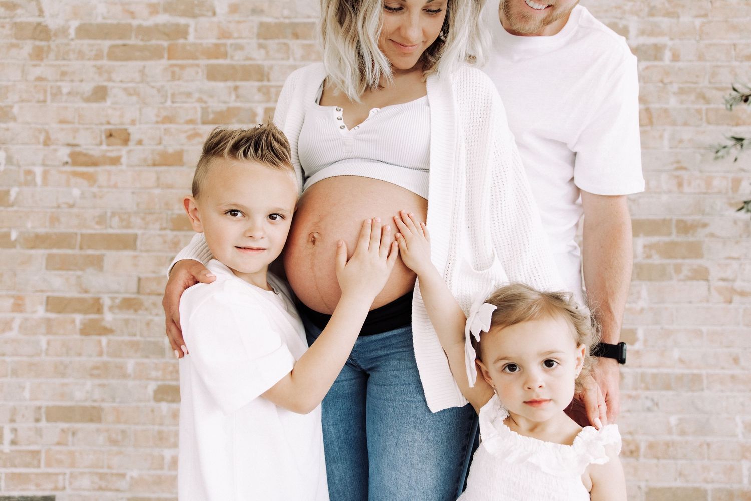 Candid family maternity photos against brick wall in casual white shirts.