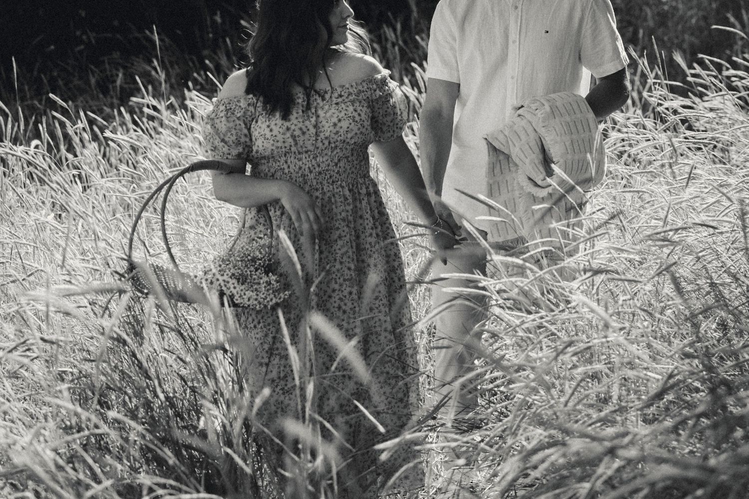 Black and white image of couple walking through tall grass field holding hands.