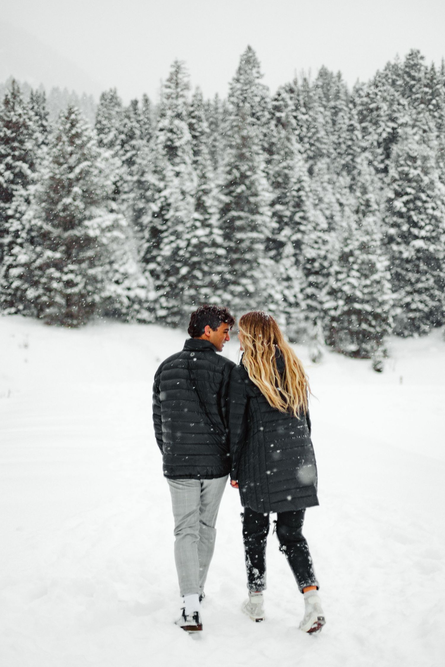Two people walk hand in hand through a snow-covered forest landscape.