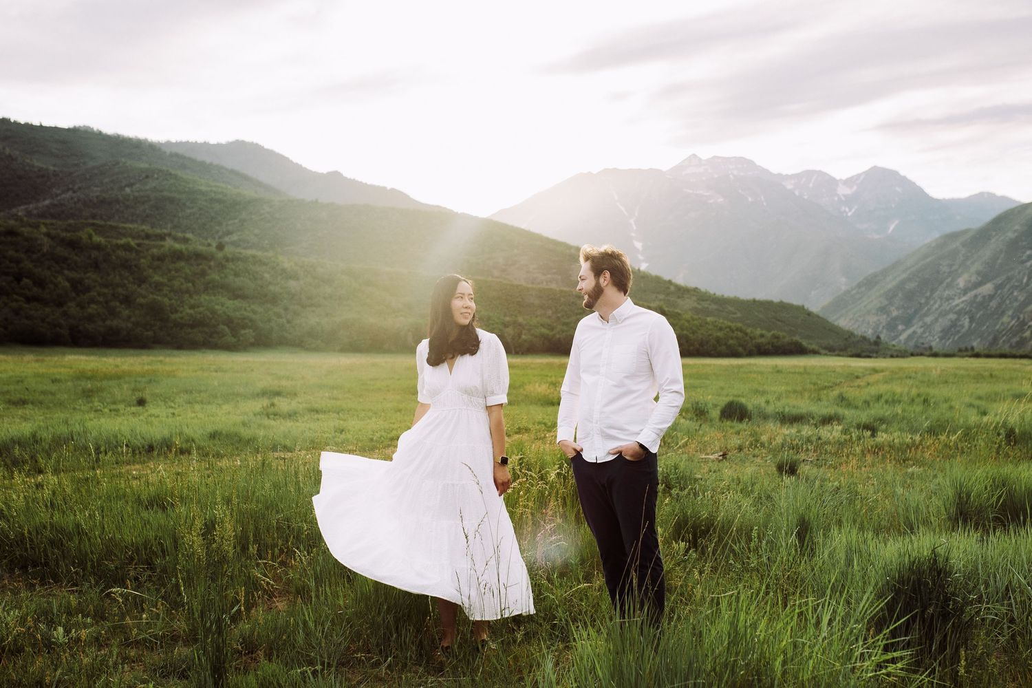 Couple in white attire standing in mountain meadow with dramatic landscape backdrop.