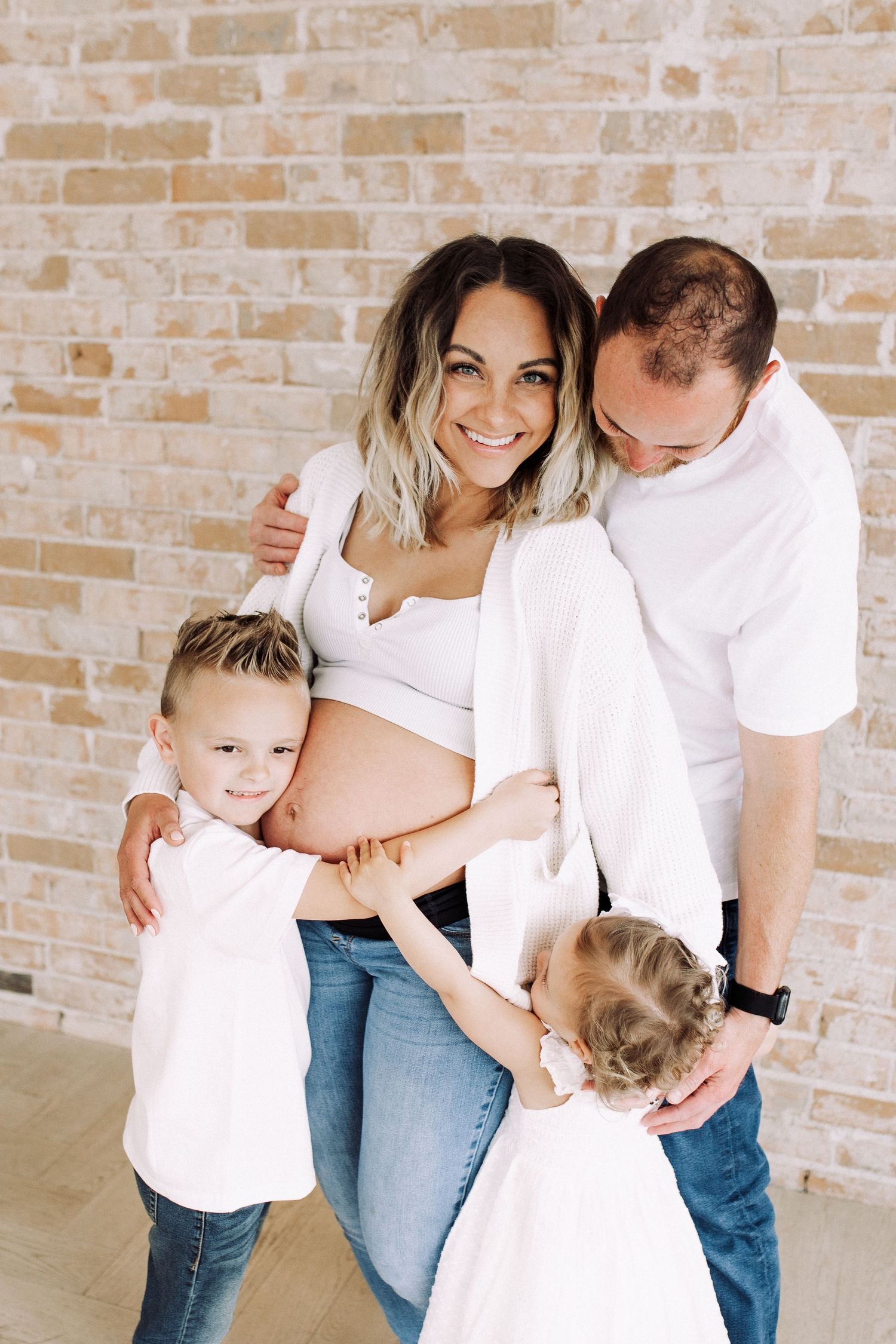 Joyful family maternity photos with everyone wearing white shirts against brick wall.