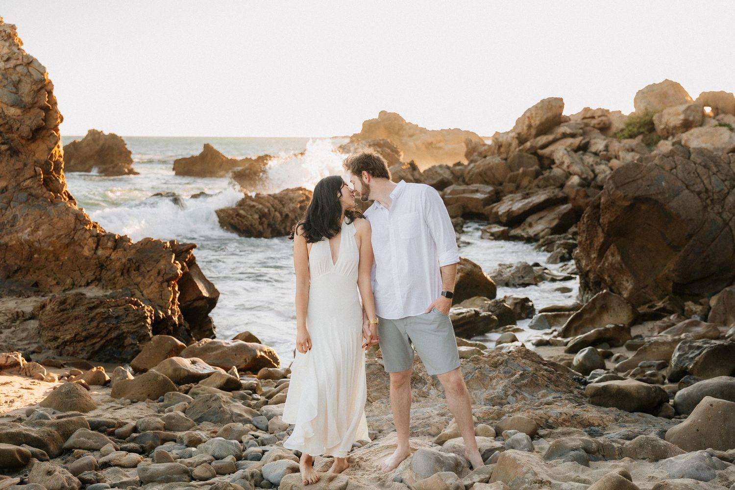 Romantic coastal photoshoot with couples walking among rocky beach formations at sunset.