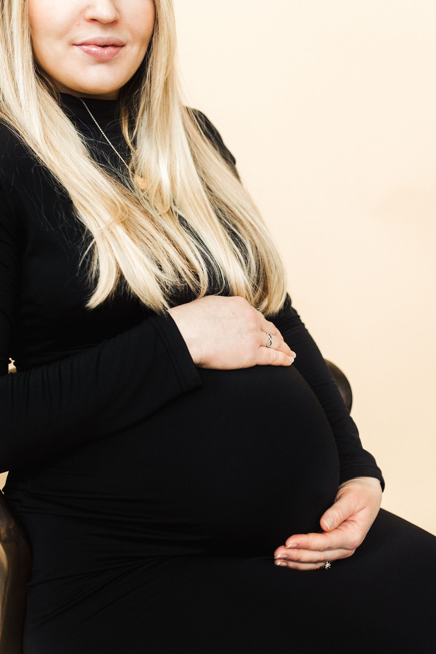 Expectant mother in black dress cradles her baby bump against cream background.