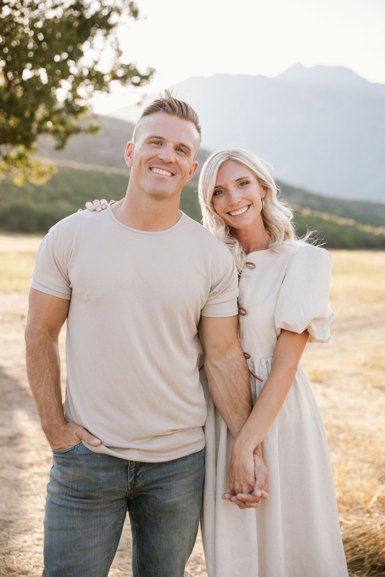 A couple poses together in casual attire against a scenic mountain backdrop during golden hour.