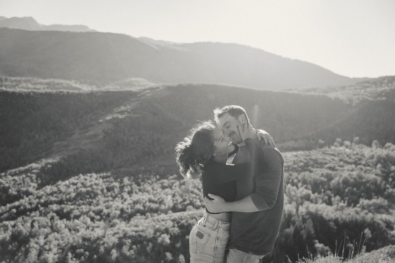 A romantic black and white portrait of a couple embracing against a mountain landscape.