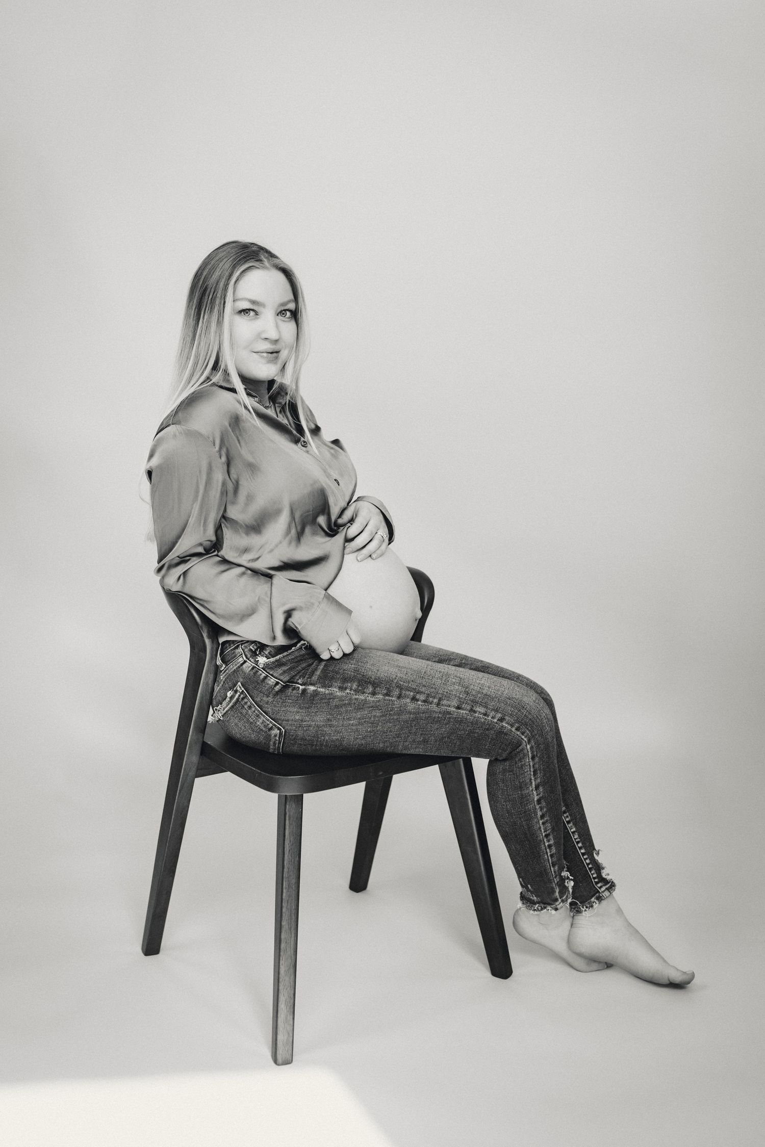 Maternity portrait in black and white of woman sitting on chair in casual attire.