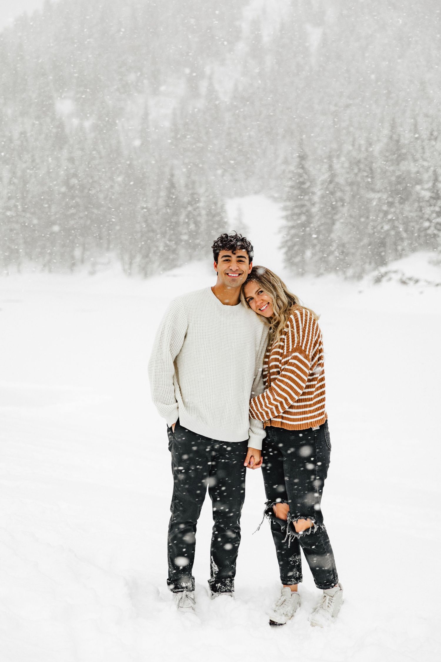 A couple embraces while standing in a snowy winter landscape with evergreen trees in the background.