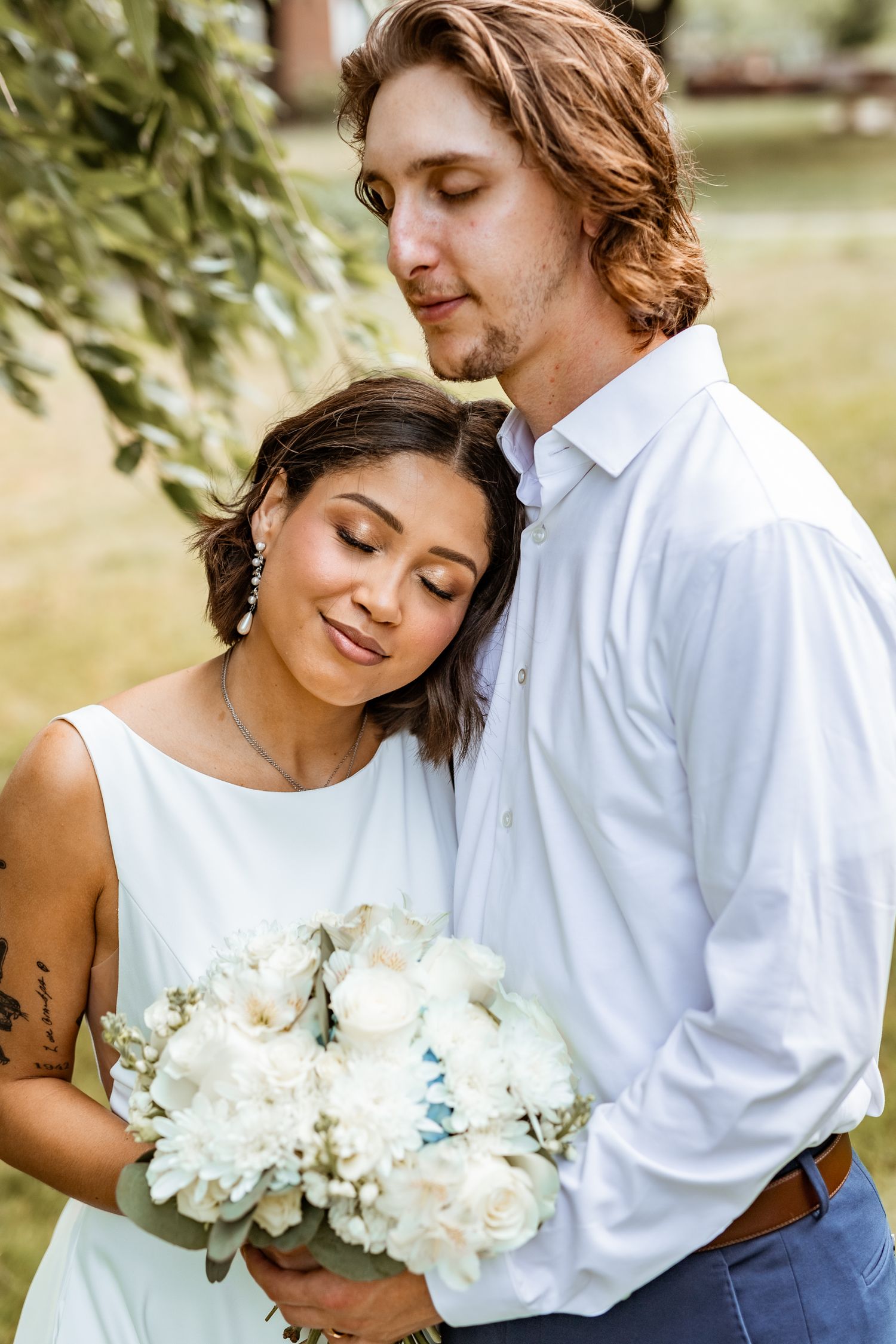 A romantic wedding couple embraces outdoors while holding a beautiful bouquet of white peonies and roses.