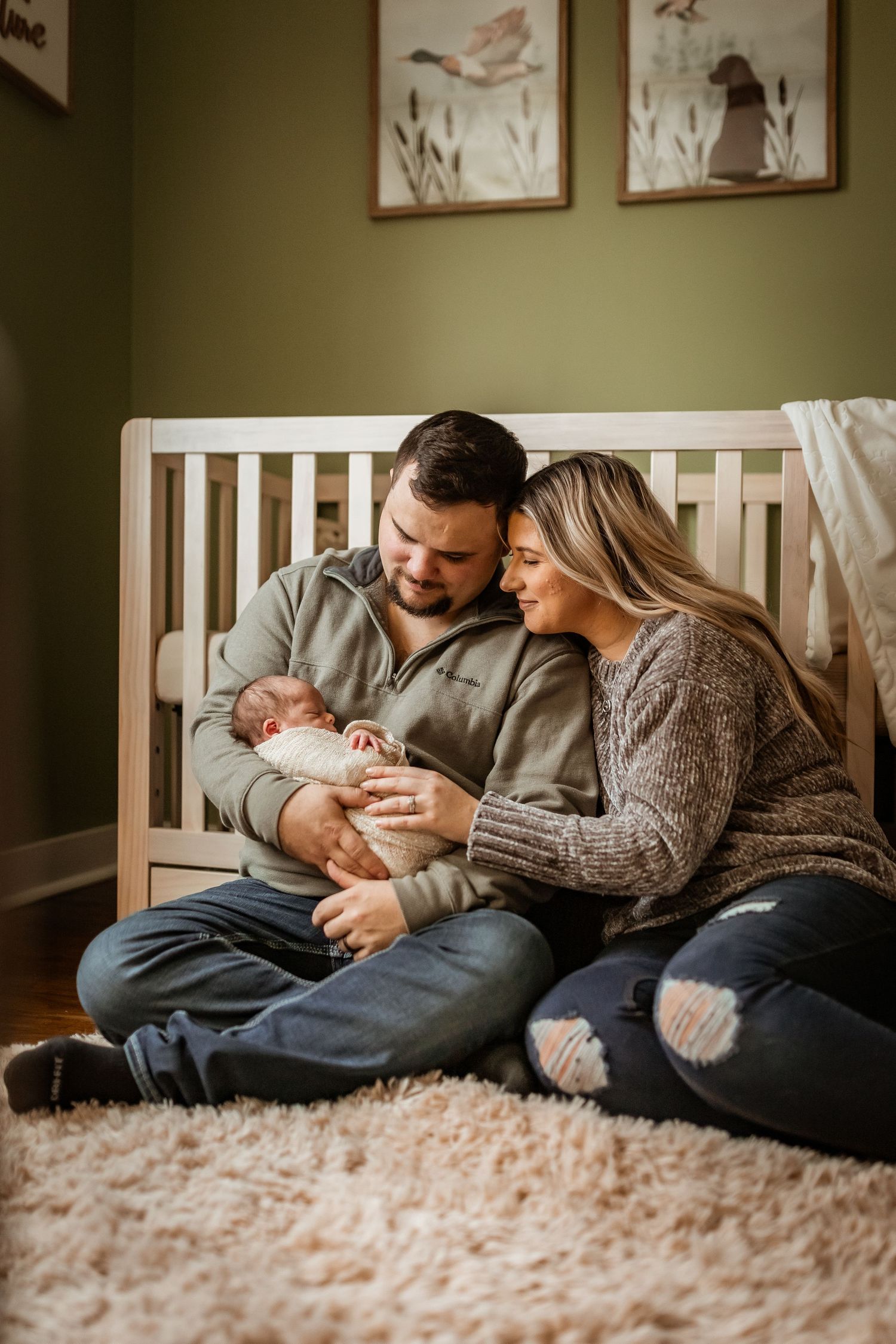 Couple sitting on floor by white crib holding newborn baby in cozy nursery with artwork on olive green walls.