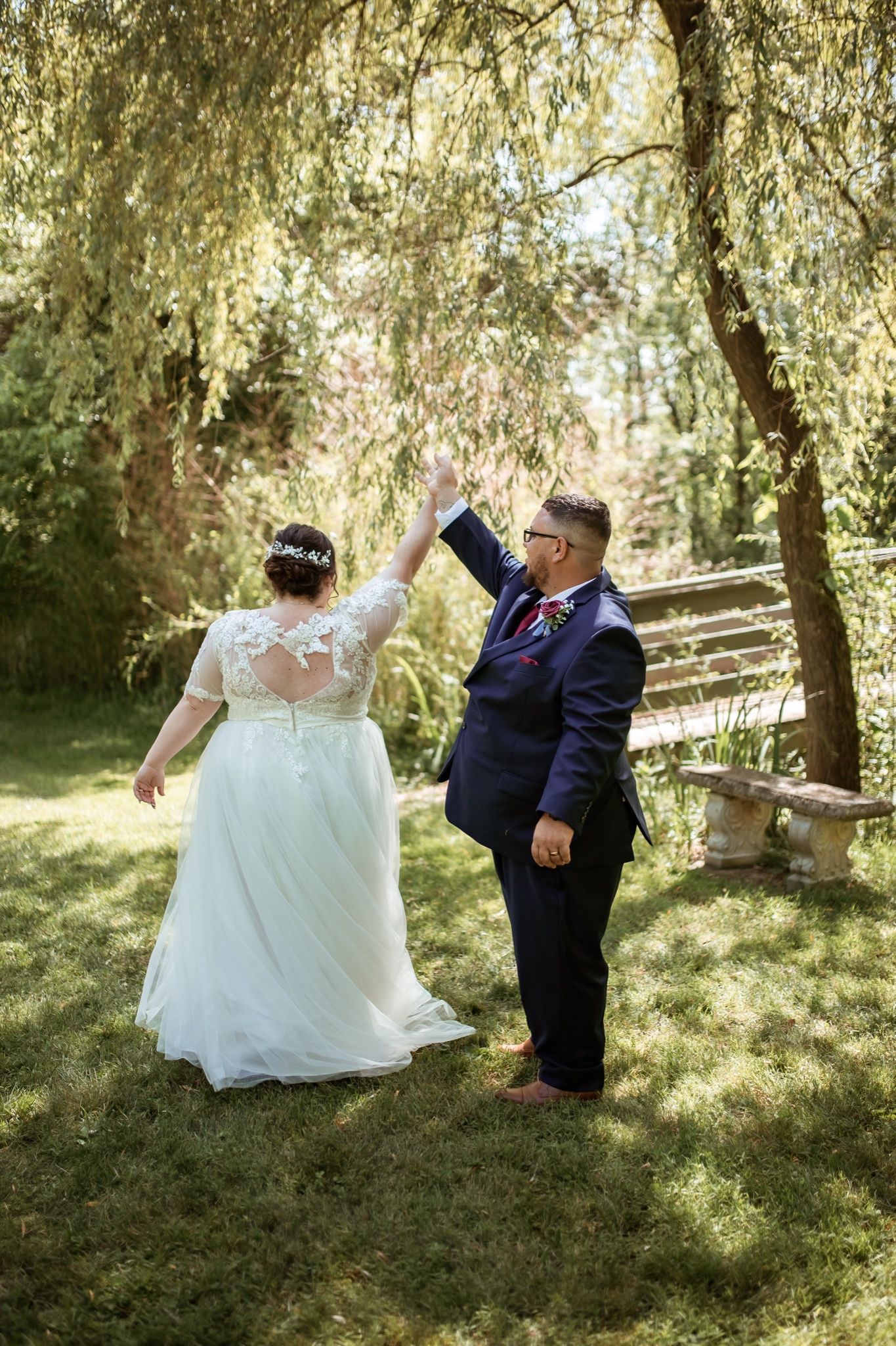 Wedding couple dancing joyfully under willow trees in a sunlit garden setting.