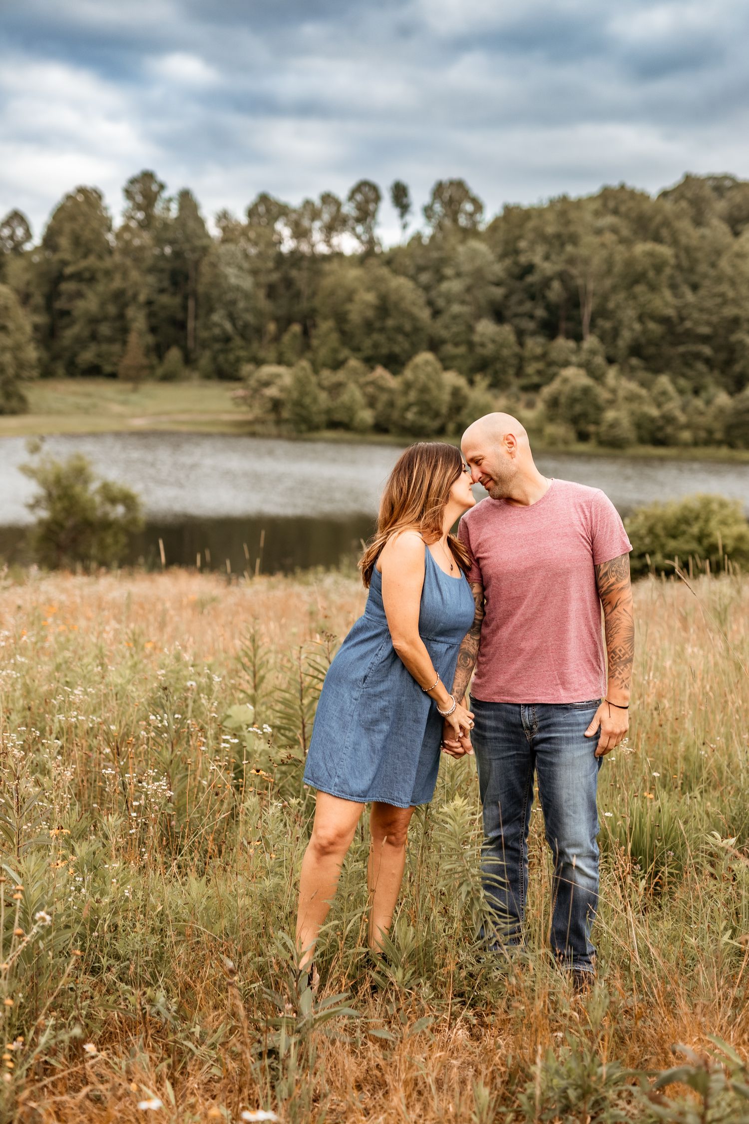 A couple shares an intimate moment in a rustic field overlooking a serene lake with trees in the background.