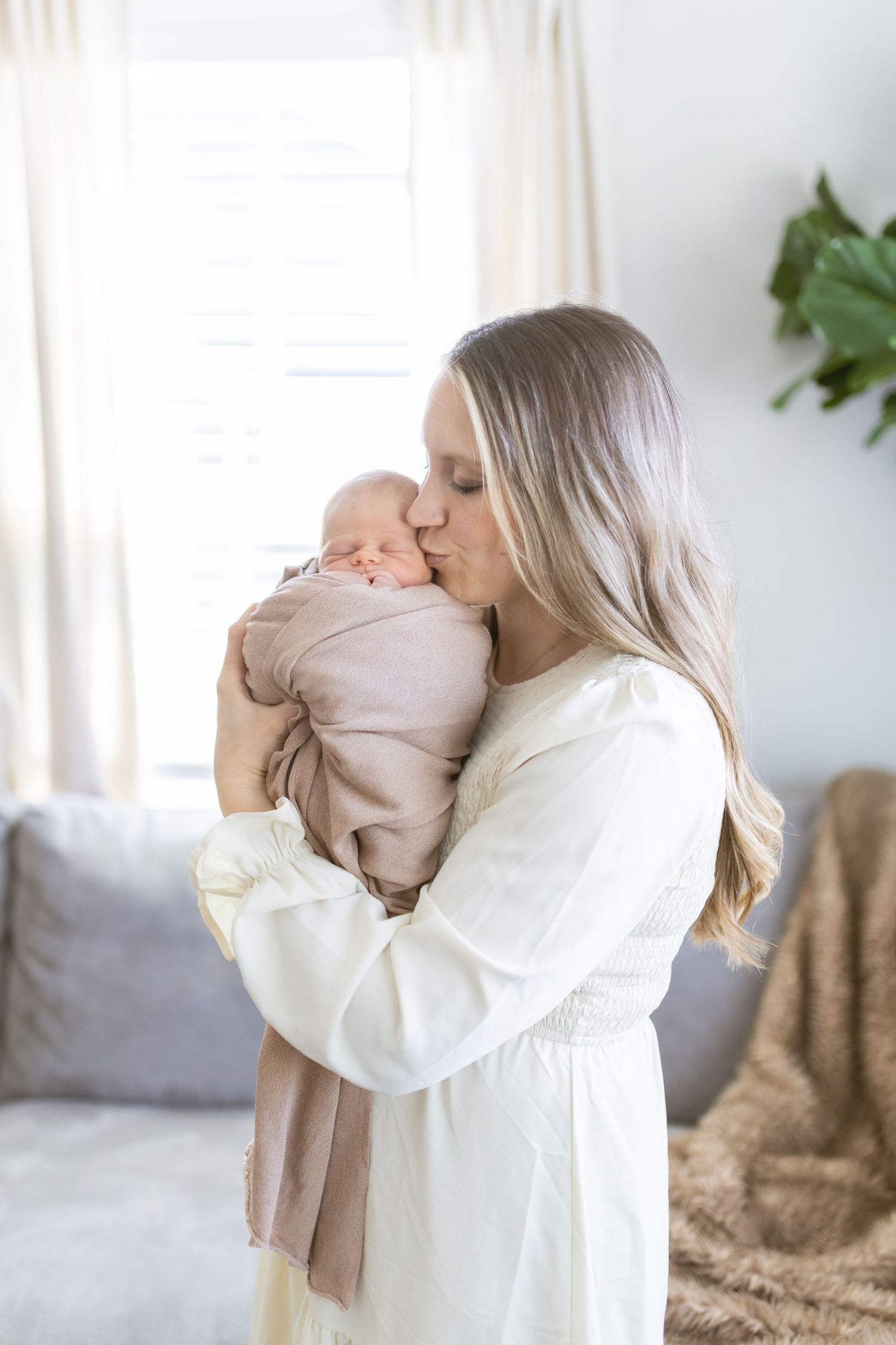 A mother in a white robe tenderly kisses her newborn baby wrapped in a pink swaddle blanket.