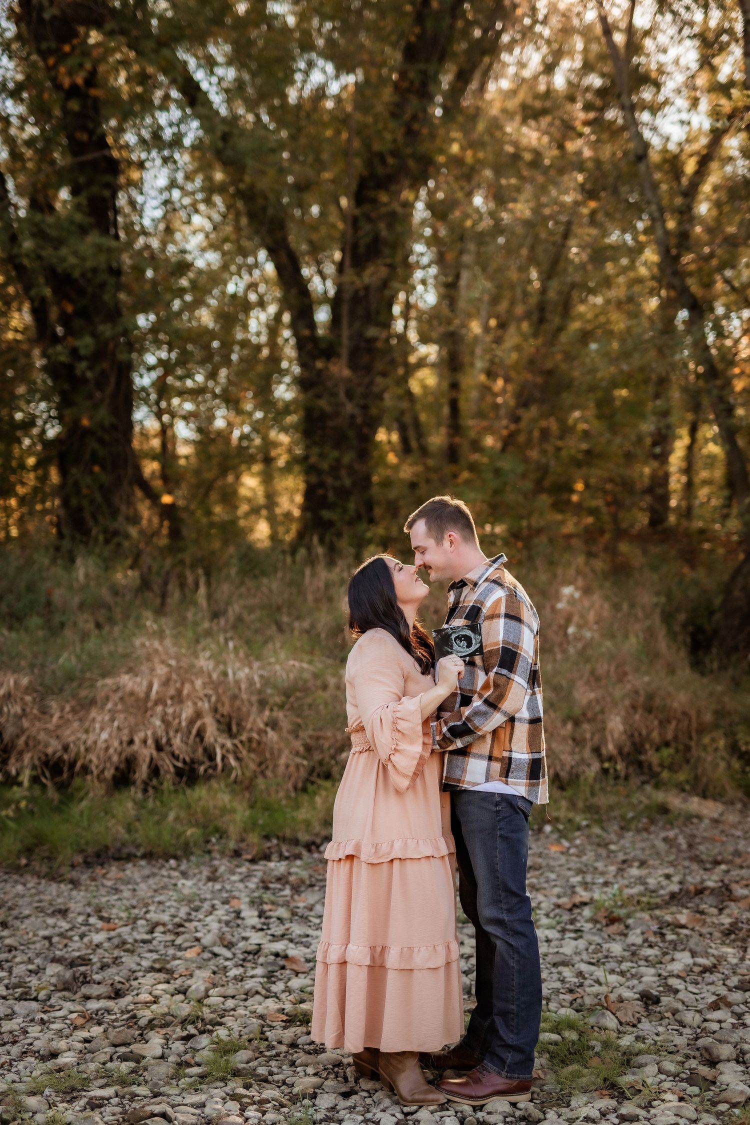 A couple in a peach dress and plaid shirt embrace on rocky ground during golden hour in a wooded setting.