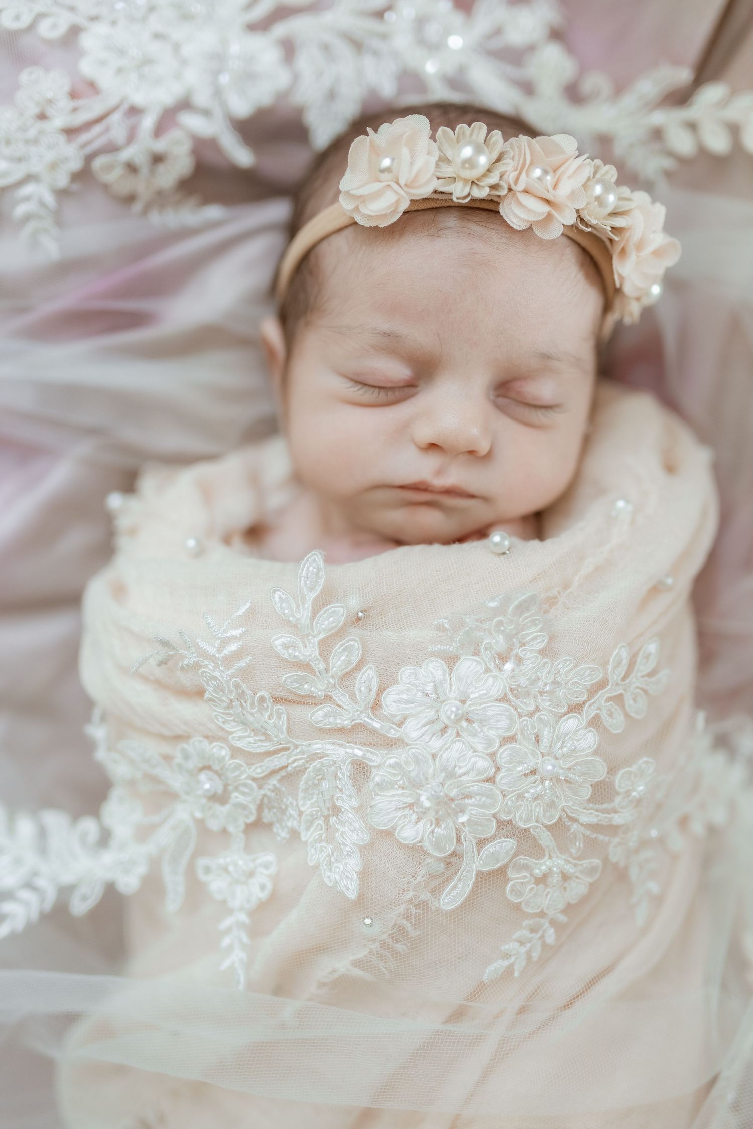 Sleeping newborn wrapped in delicate white lace with an ornate headpiece on light fabric.