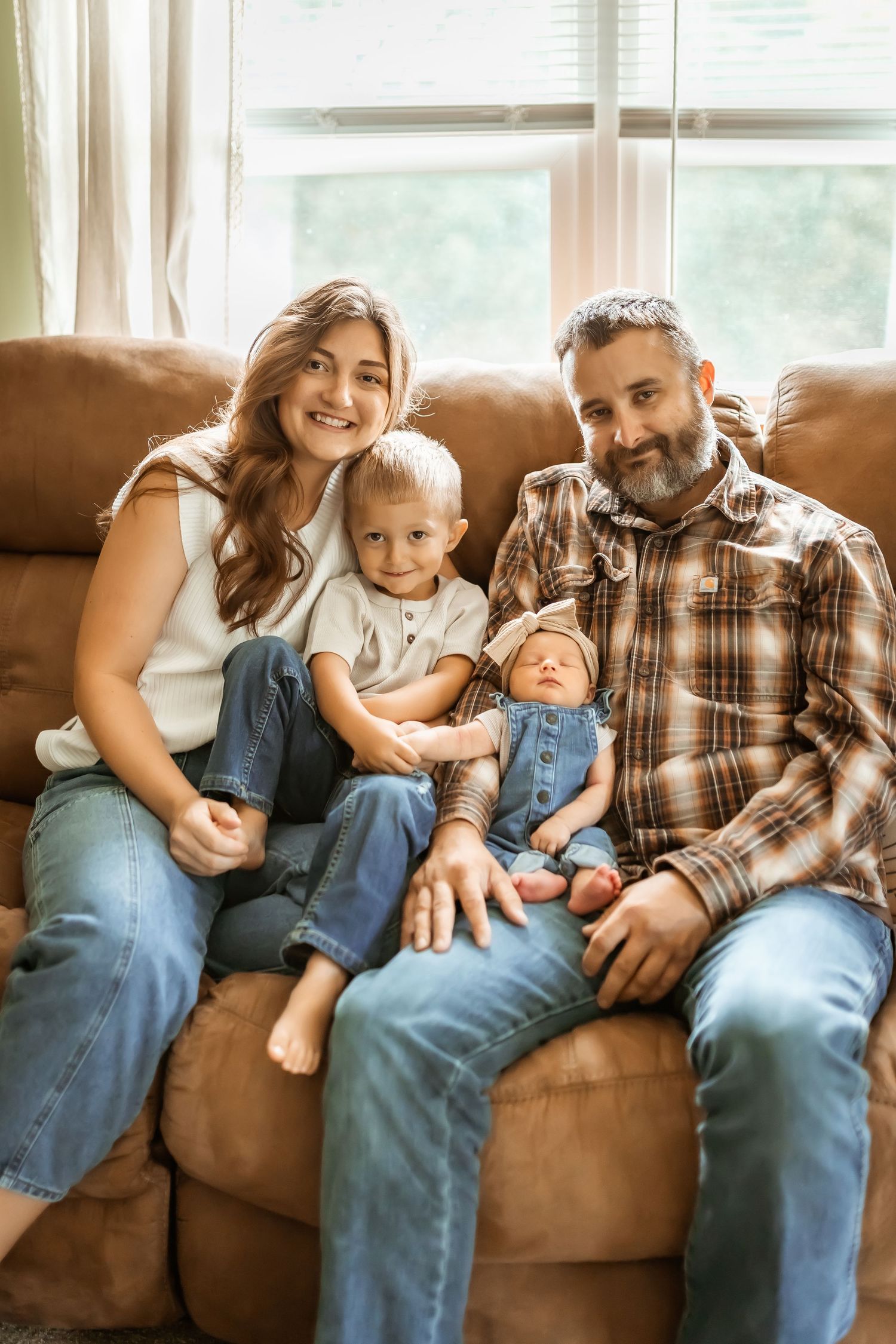 Family relaxes together on brown couch in living room with natural window light.