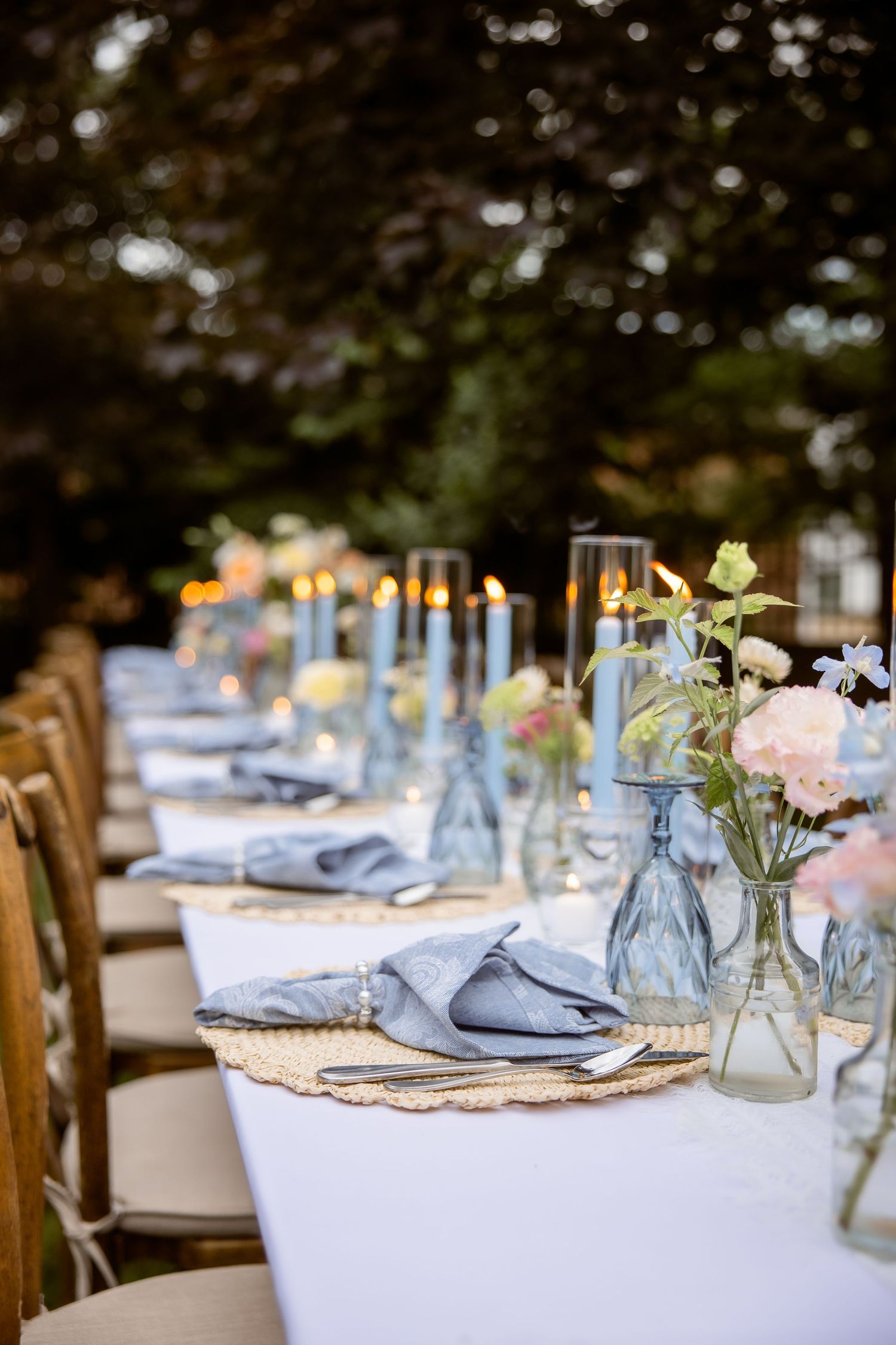 Elegant outdoor wedding reception table set with blue candles, floral arrangements, gray linen napkins, and glassware at dusk.