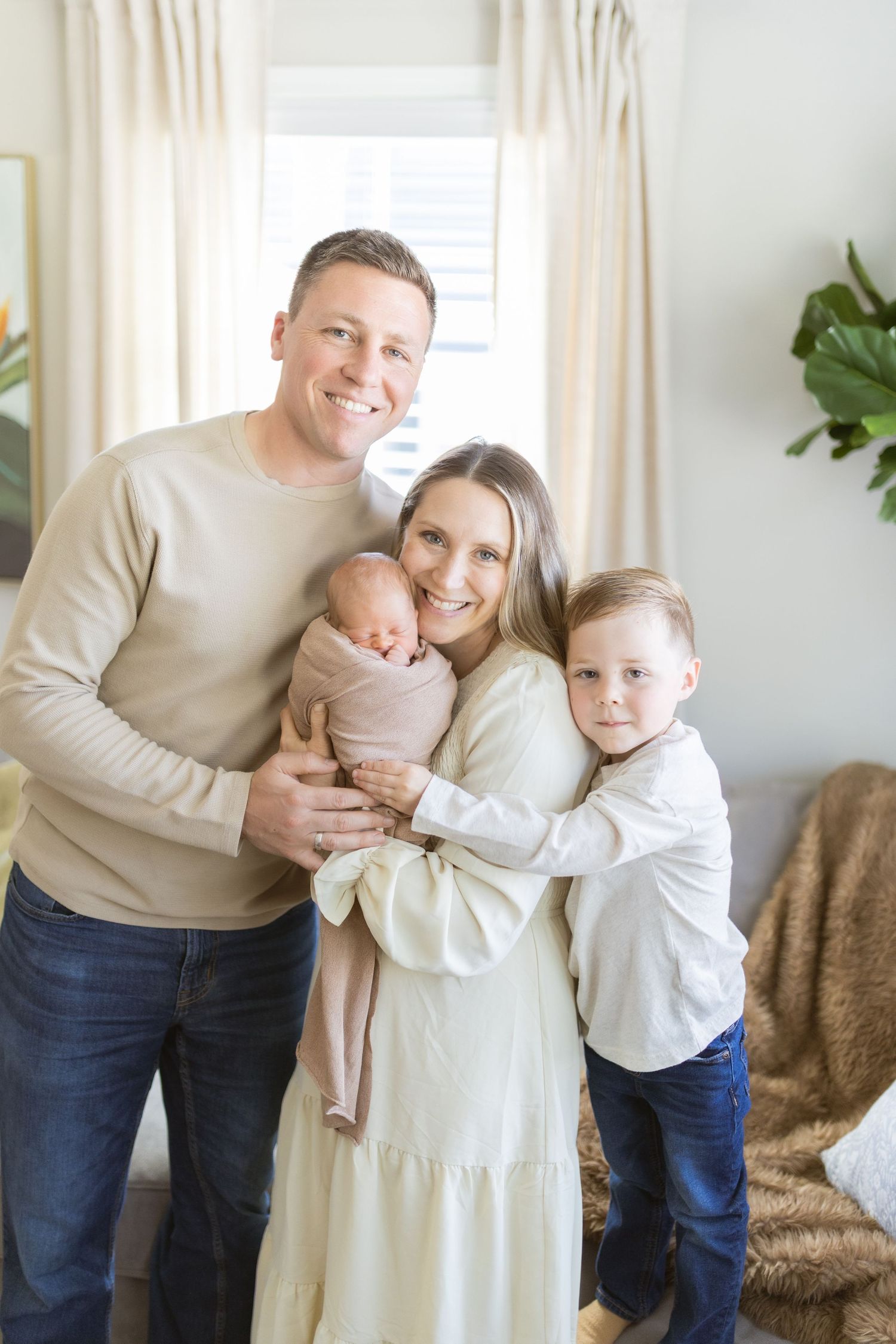 Happy family of four posing indoors, parents smiling while mom holds newborn baby, young son hugging mom in bright living room.