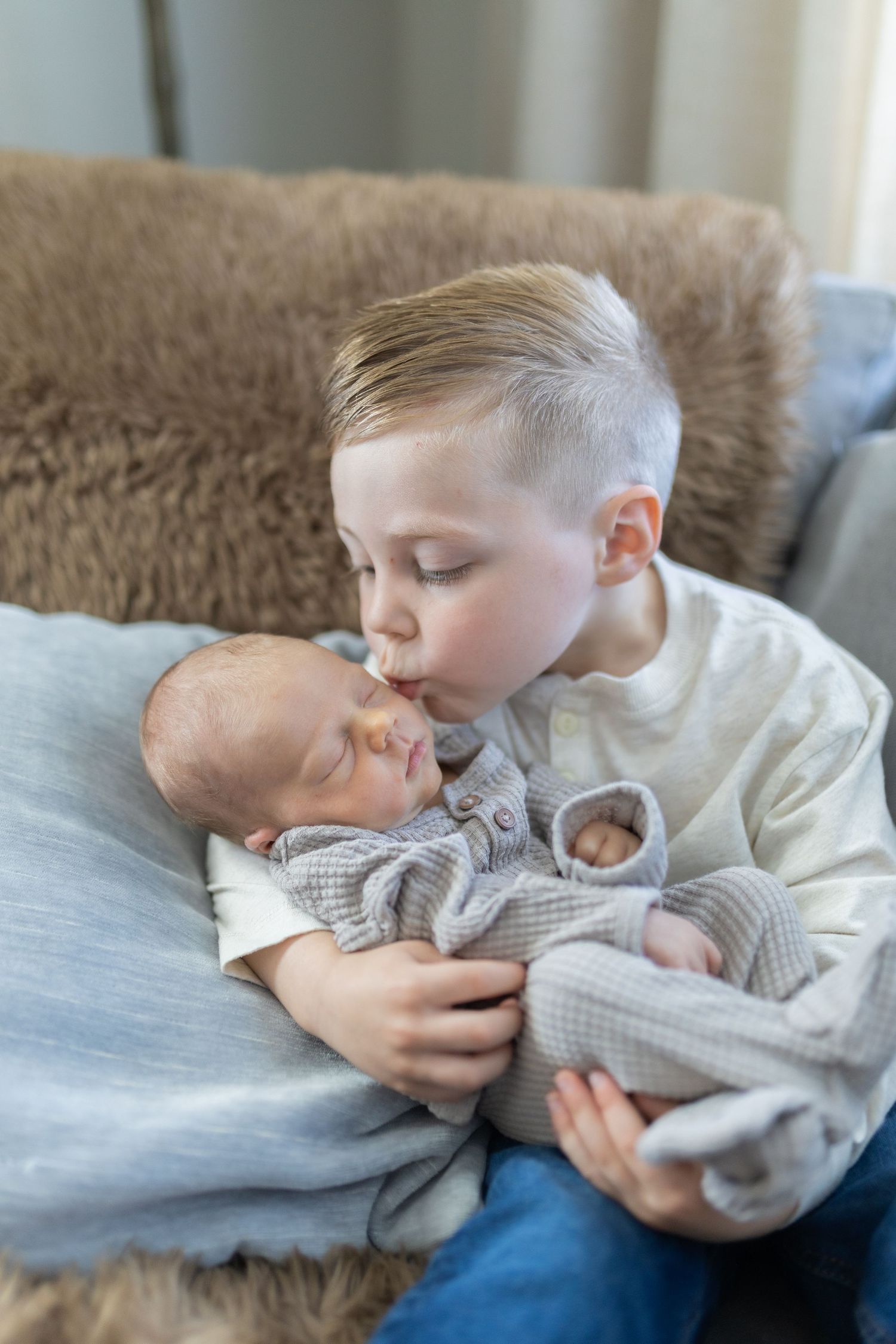Young boy gently kissing sleeping newborn baby sibling while holding them on a cozy sofa.