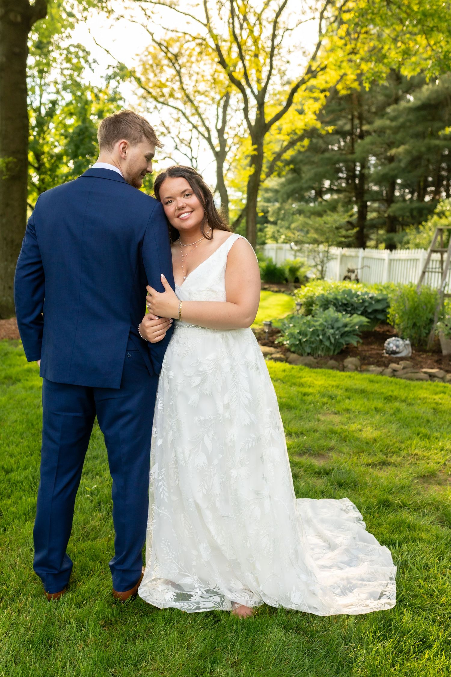 Bride in white lace gown smiles at groom in navy suit during outdoor wedding portrait in lush green garden.
