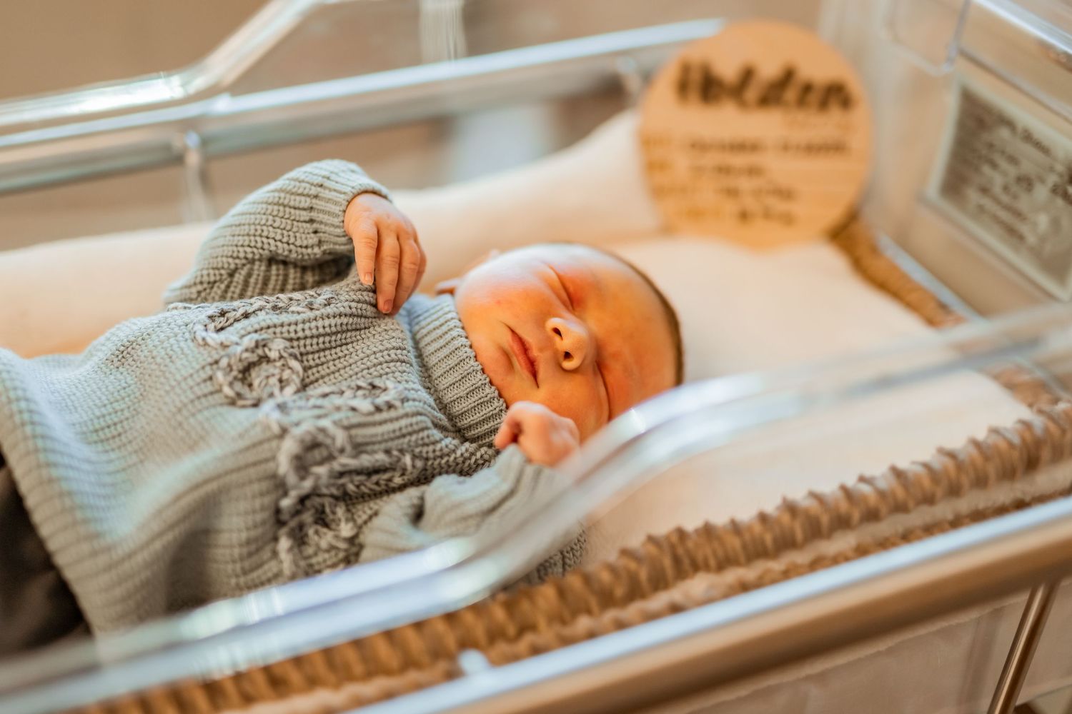 Newborn baby wearing a gray knit sweater rests peacefully in hospital bassinet.
