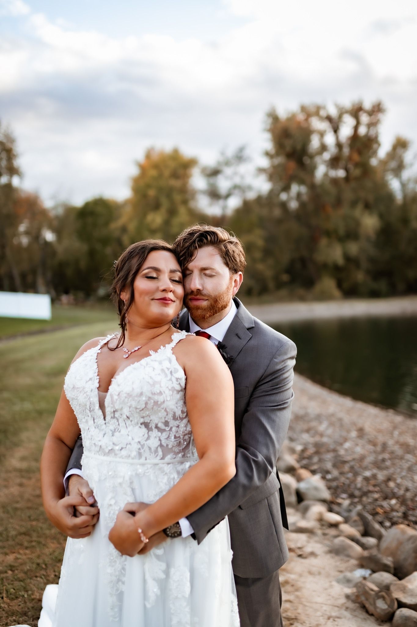 A couple in wedding attire embrace by a lake with fall trees in the background on their special day.