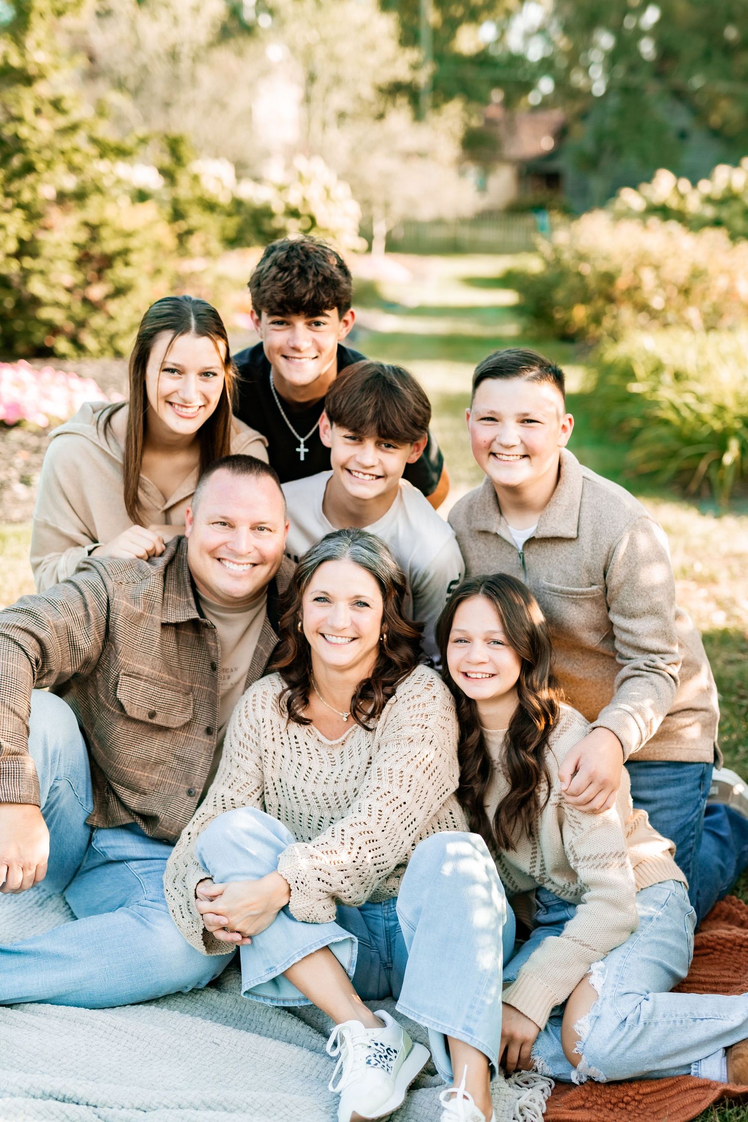 A family group sits together outdoors on a blanket in a garden setting with natural lighting and casual attire.