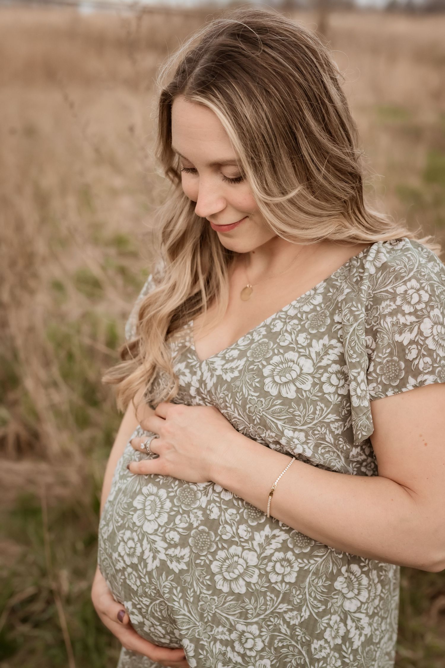 Pregnant woman in floral sage dress cradling baby bump in golden field, smiling softly downward.