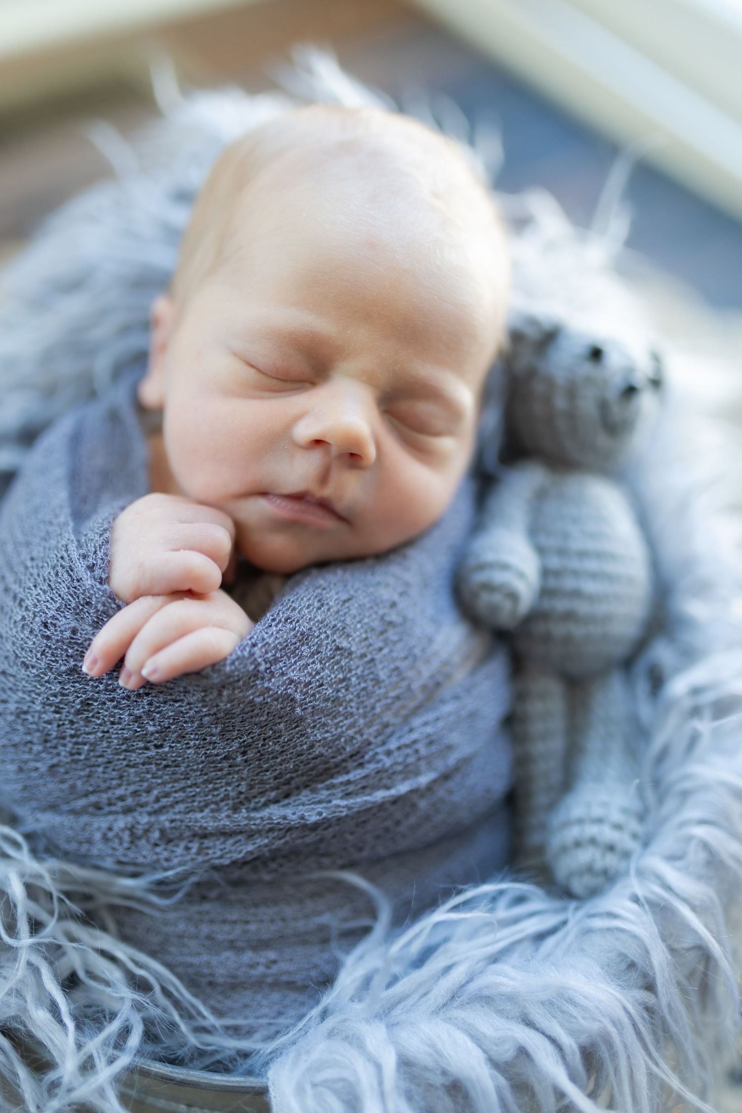 Newborn baby sleeping peacefully wrapped in gray knit blanket beside a small crocheted teddy bear toy.