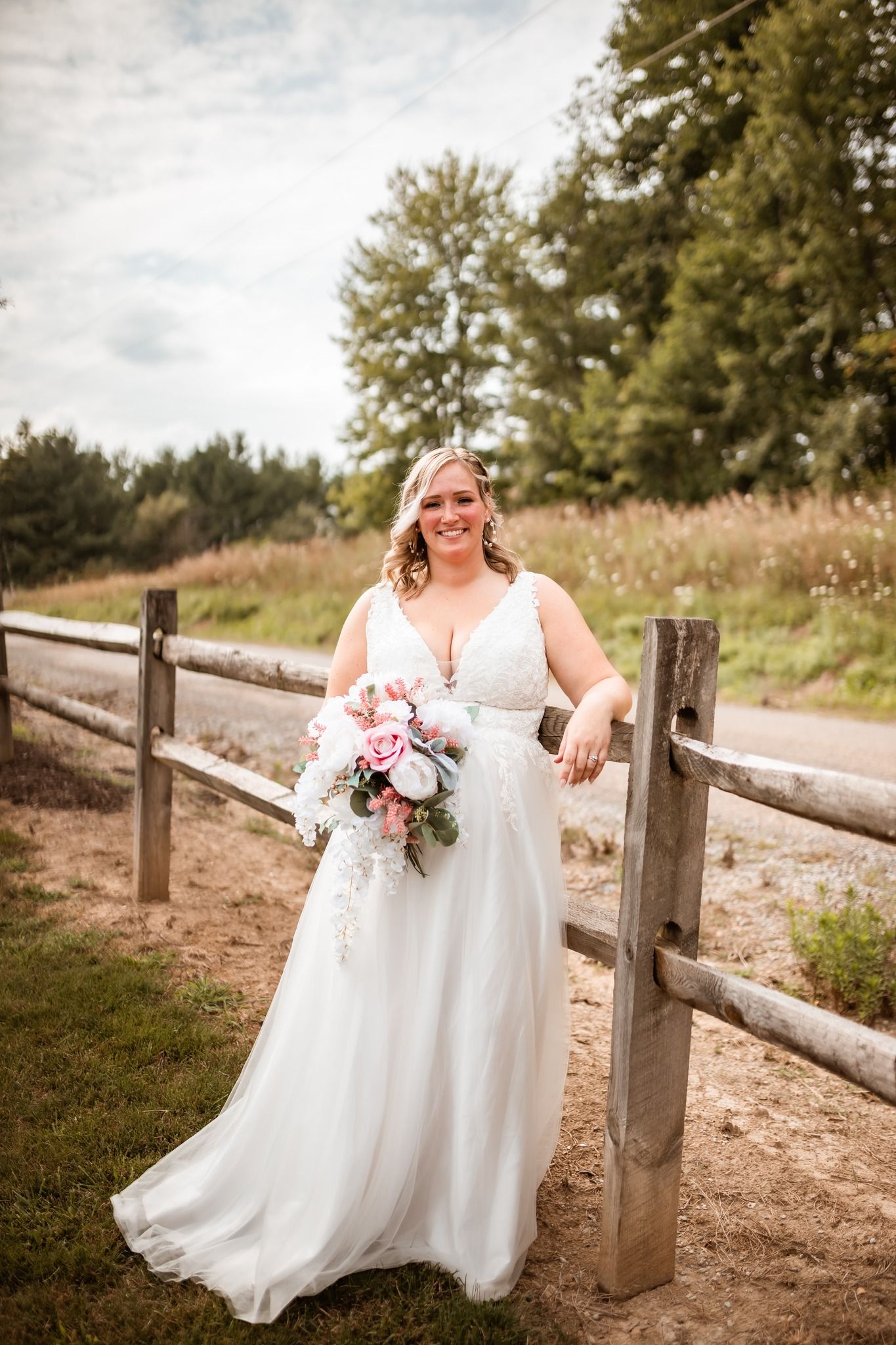 A bridal portrait shows a joyful moment on a rustic path with wooden fencing and a lush bouquet of pink flowers.