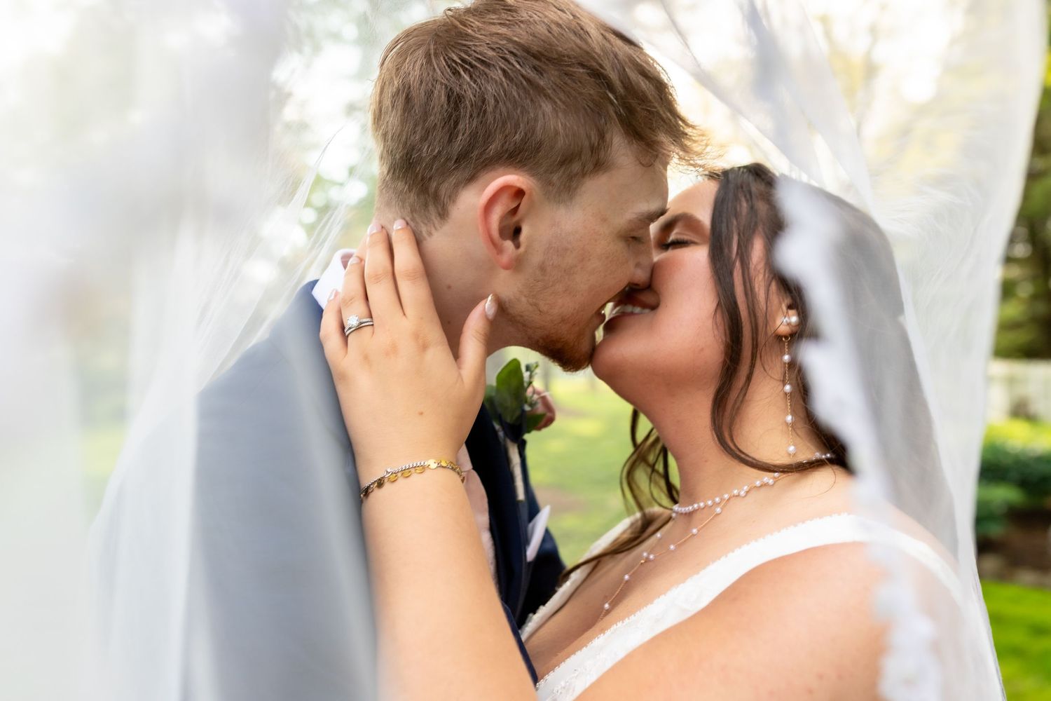 Bride and groom share a romantic kiss under a flowing white veil outdoors on their wedding day.